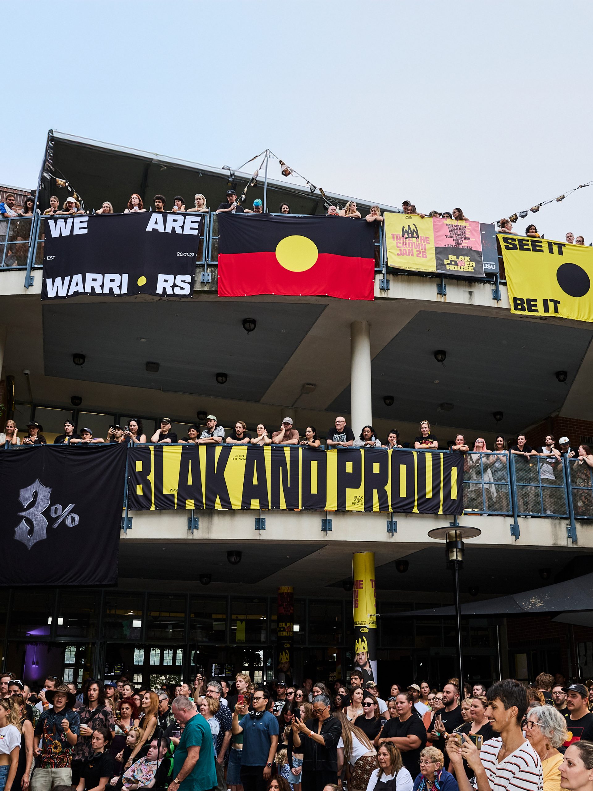 A photo looking up to a two level building, crowds filling the balconies. Bright posters and the Aboriginal flag hangs over the edges, while the crowd looks to the left watching a stage show.