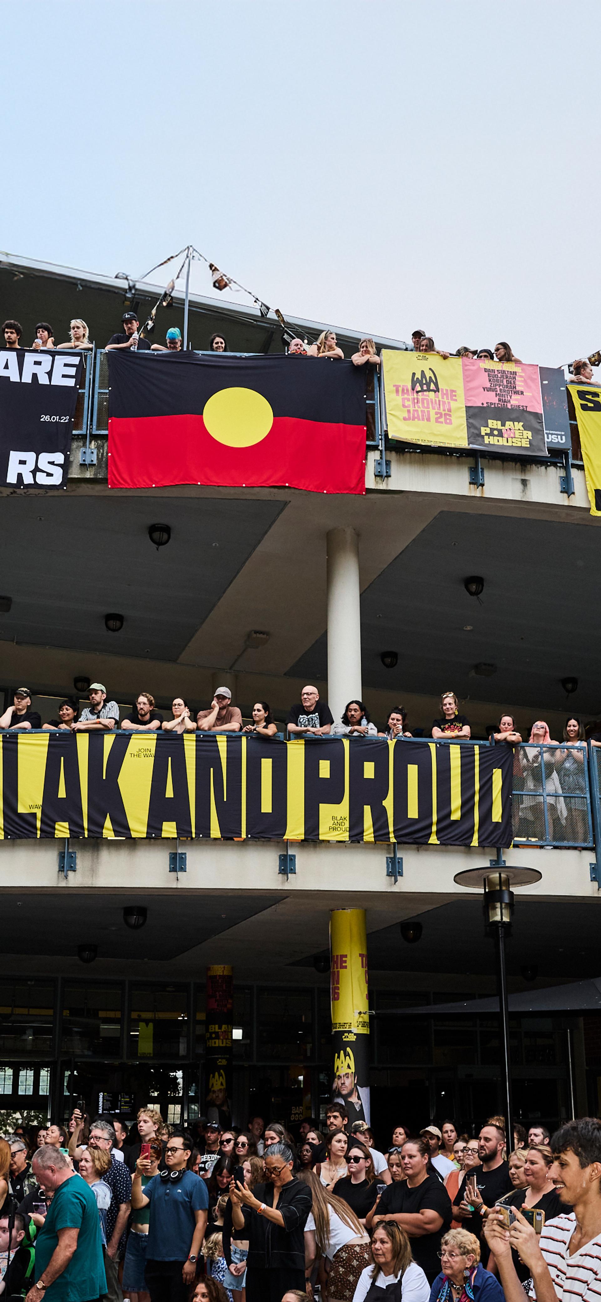 A photo looking up to a two level building, crowds filling the balconies. Bright posters and the Aboriginal flag hangs over the edges, while the crowd looks to the left watching a stage show.