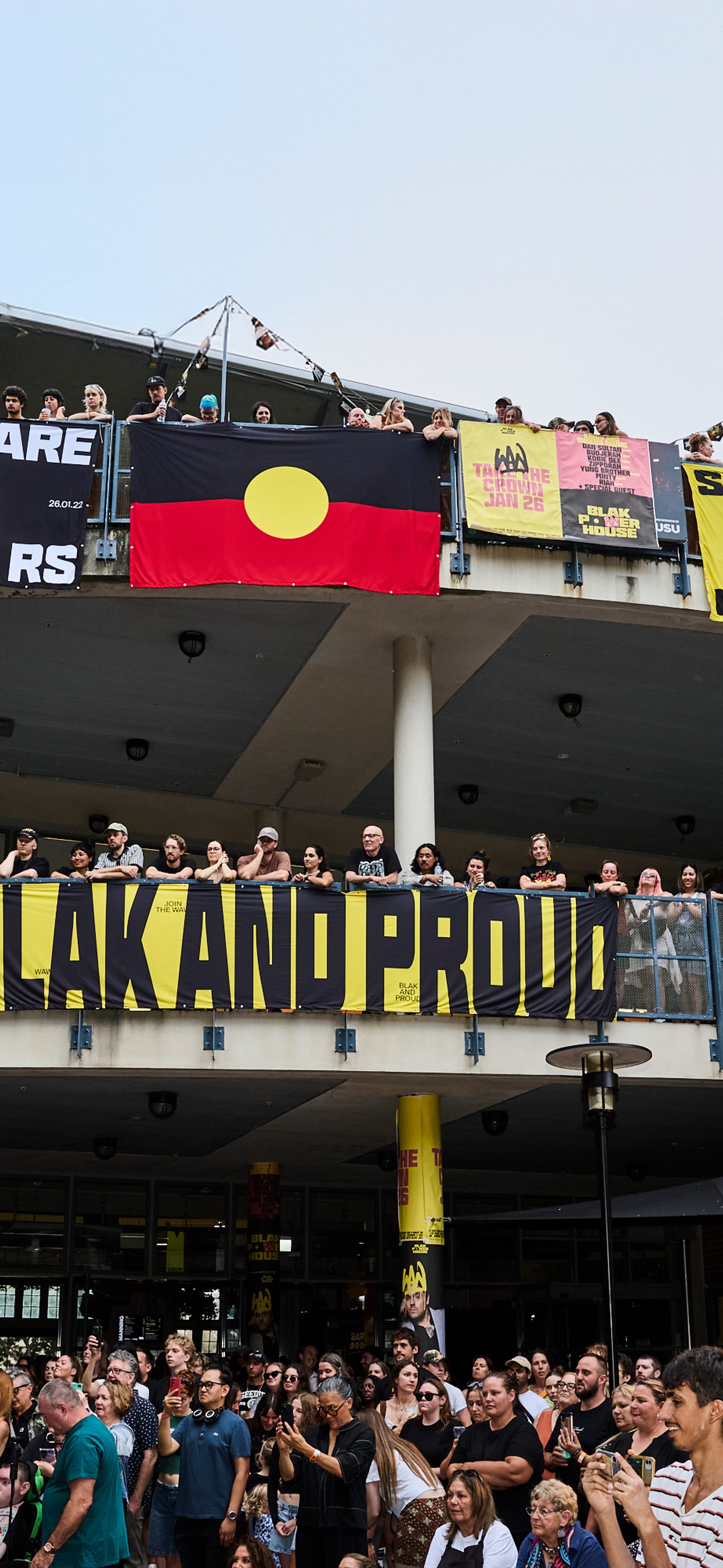 A photo looking up to a two level building, crowds filling the balconies. Bright posters and the Aboriginal flag hangs over the edges, while the crowd looks to the left watching a stage show.