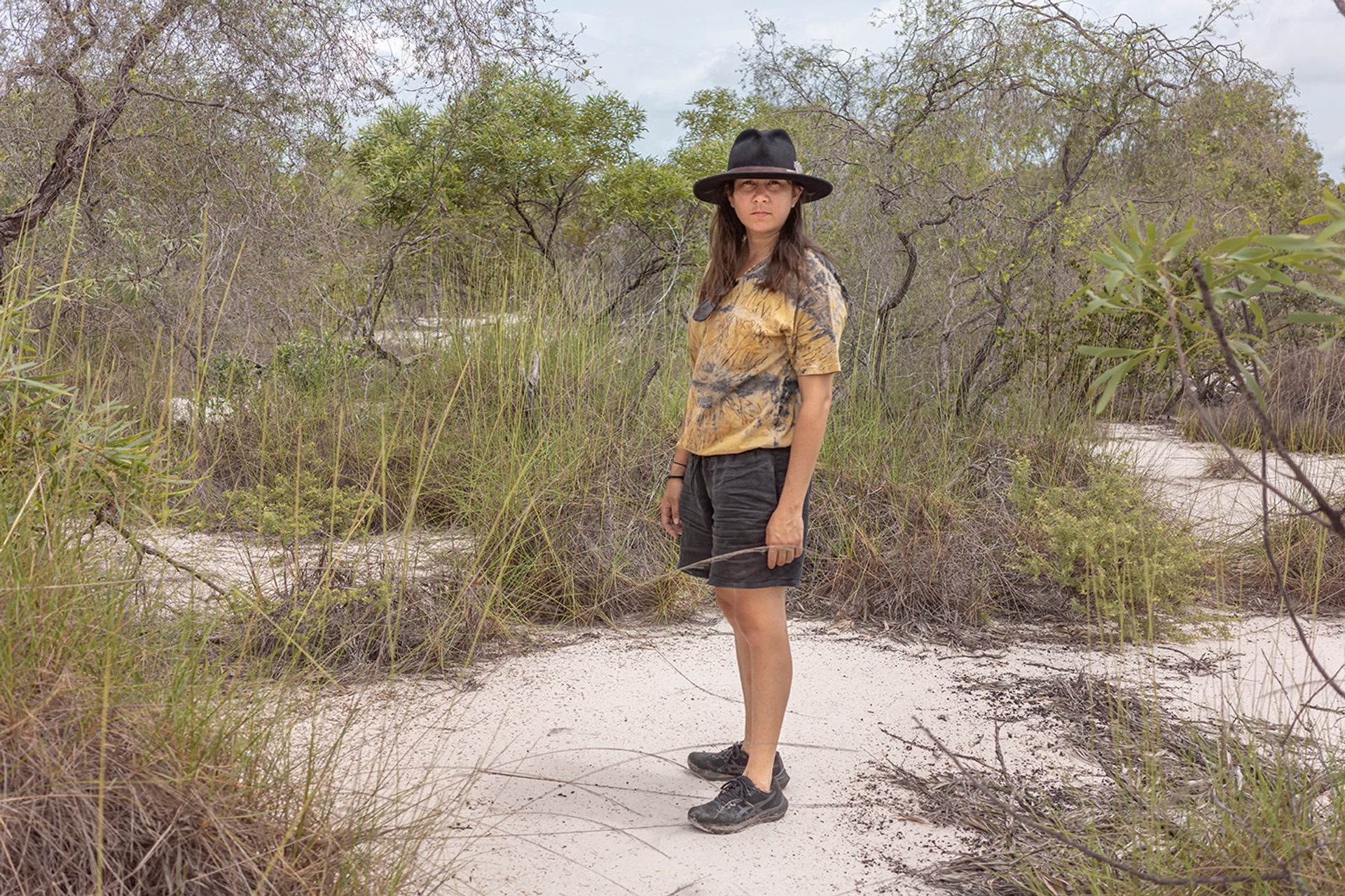 A coastal landscape with flat white sand, green trees, patches of long green grass. A woman stands in the middle of environment, on a clearing of sand. Her body is in profile and her face looks directly at the camera.
