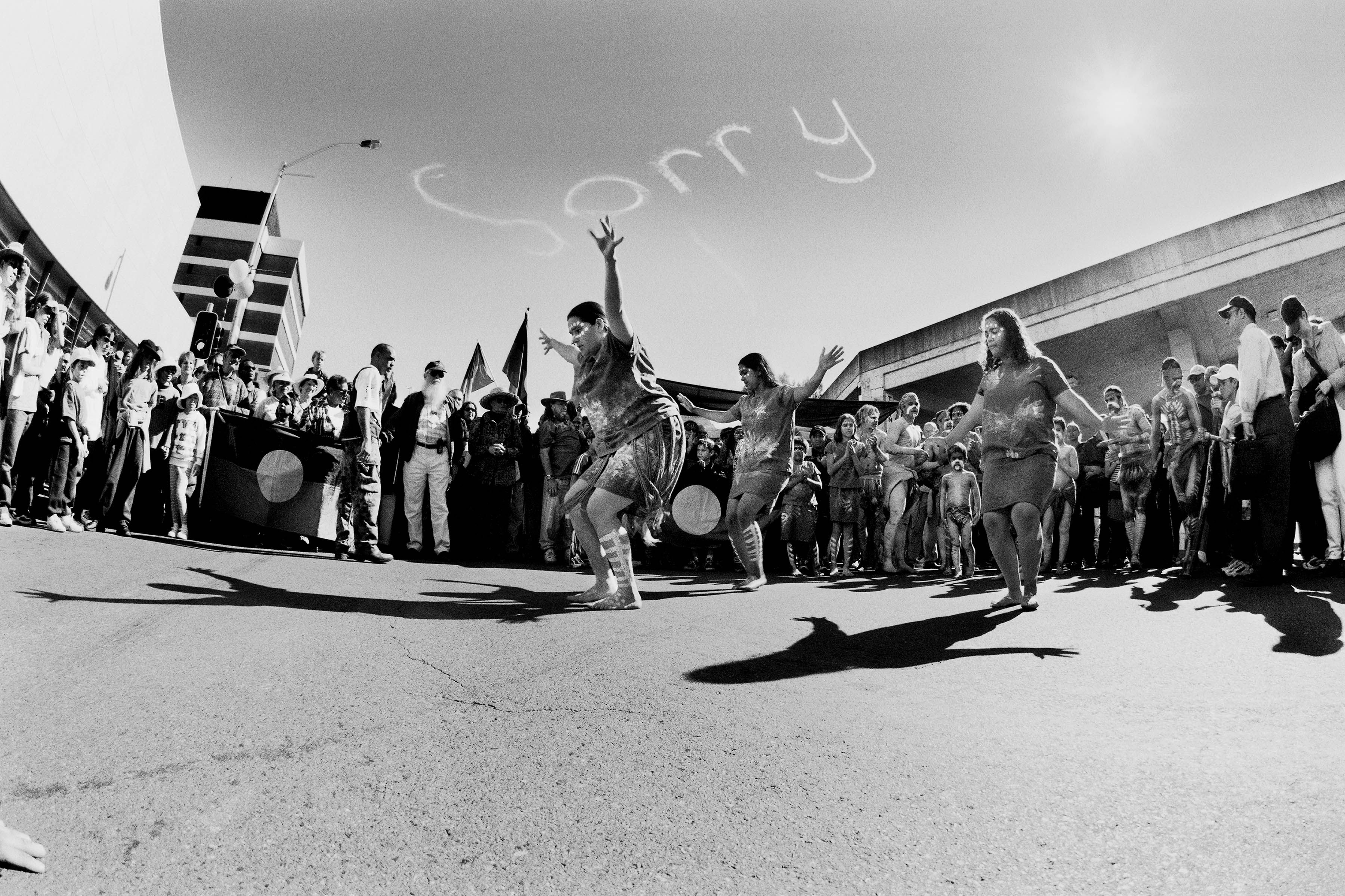 Australian First Nations people dancing underneath a sky saying 'Sorry'