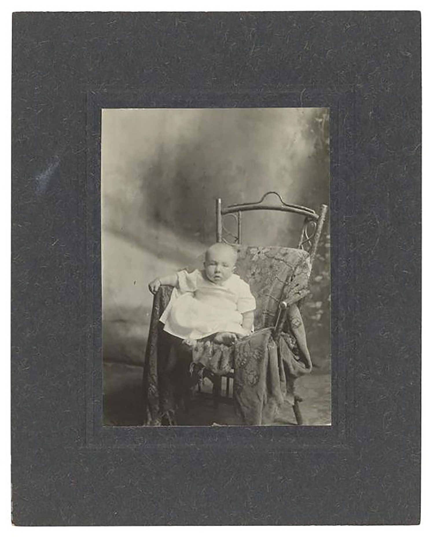 Full length studio portrait of John Hollis Pearce aged about 1 year. He is posed on a chair with tapestry blanket and dressed in a white smocking gown, looking at the camera.