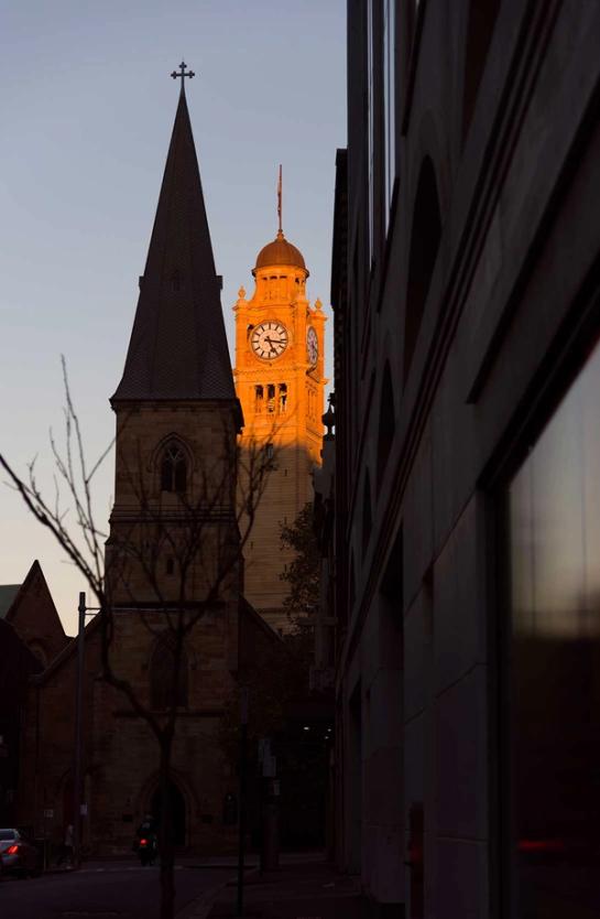 A clock on a tall building illuminated by the sunset through two shaded buildings.