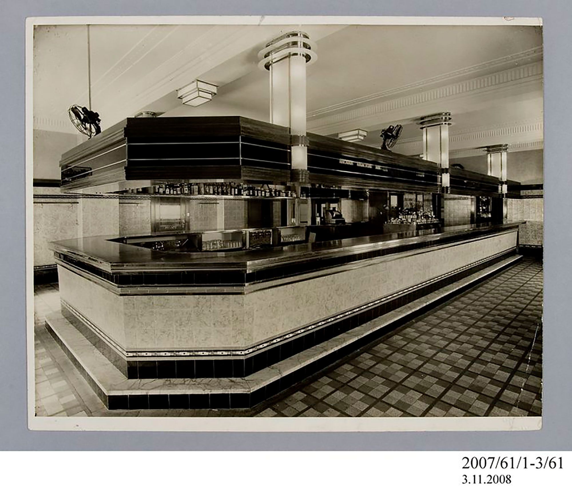 Photographic print, black & white, private dining room in Henson Park Hotel, Marrickville. In the room is a buffet, a rectangular dining table with two seats, a fireplace, a mirror and plants. On the dining table is a crystal vase with white flowers in it. The mirror is positioned above the large fireplace; there is a smaller vase with flowers on top of it. There are a few glasses and more flowers on the buffet table. The floor is carpeted with an abstract pattern.