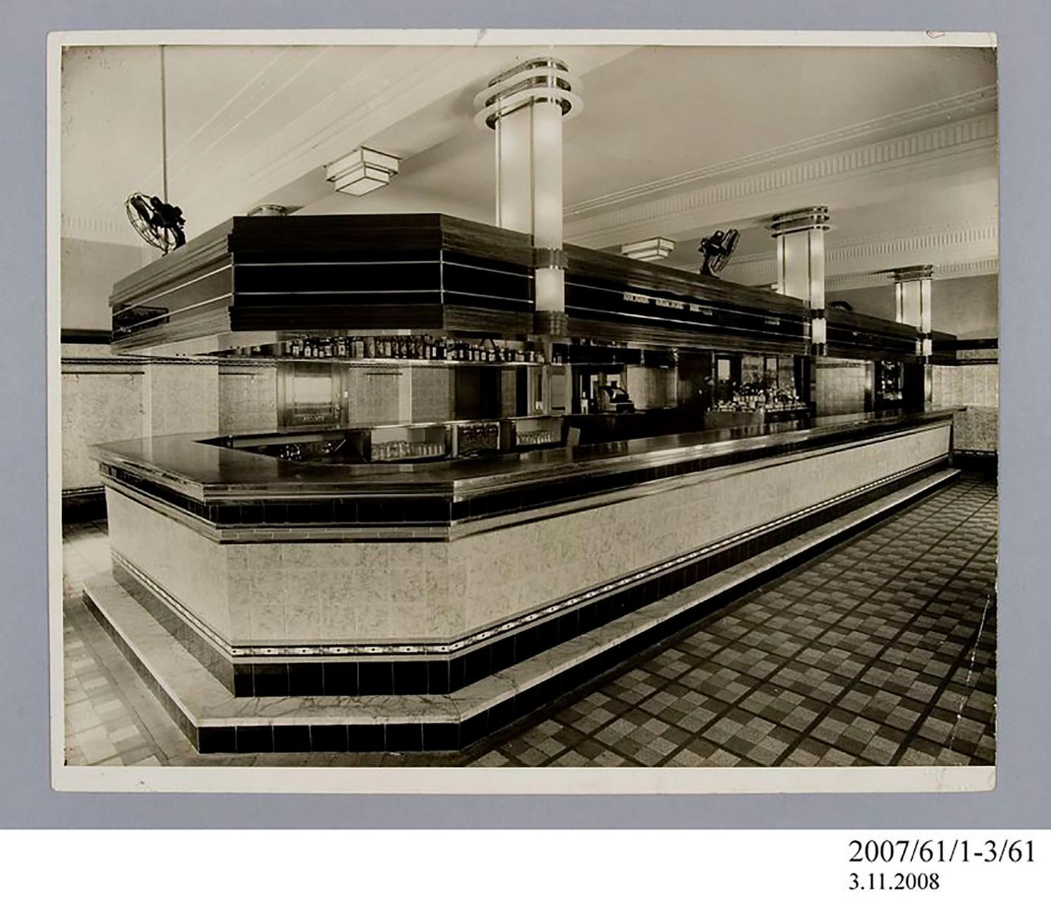 Photographic print, black & white, private dining room in Henson Park Hotel, Marrickville. In the room is a buffet, a rectangular dining table with two seats, a fireplace, a mirror and plants. On the dining table is a crystal vase with white flowers in it. The mirror is positioned above the large fireplace; there is a smaller vase with flowers on top of it. There are a few glasses and more flowers on the buffet table. The floor is carpeted with an abstract pattern.