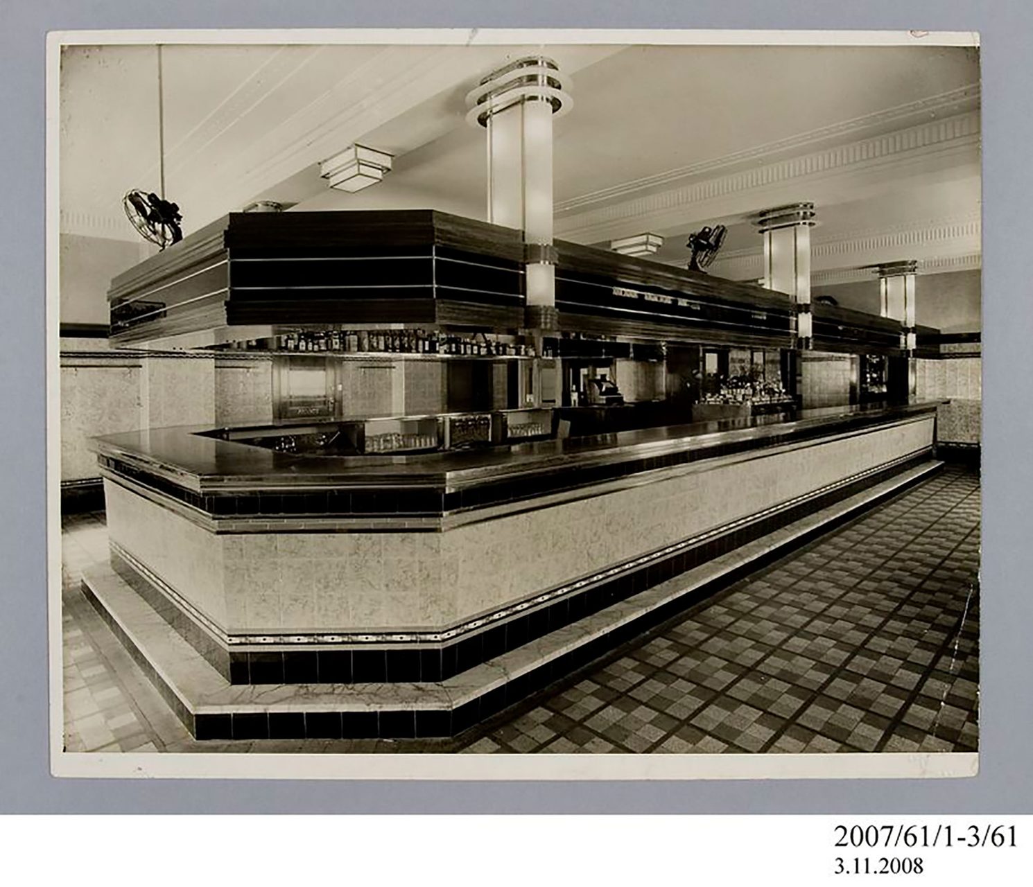 Photographic print, black & white, private dining room in Henson Park Hotel, Marrickville. In the room is a buffet, a rectangular dining table with two seats, a fireplace, a mirror and plants. On the dining table is a crystal vase with white flowers in it. The mirror is positioned above the large fireplace; there is a smaller vase with flowers on top of it. There are a few glasses and more flowers on the buffet table. The floor is carpeted with an abstract pattern.