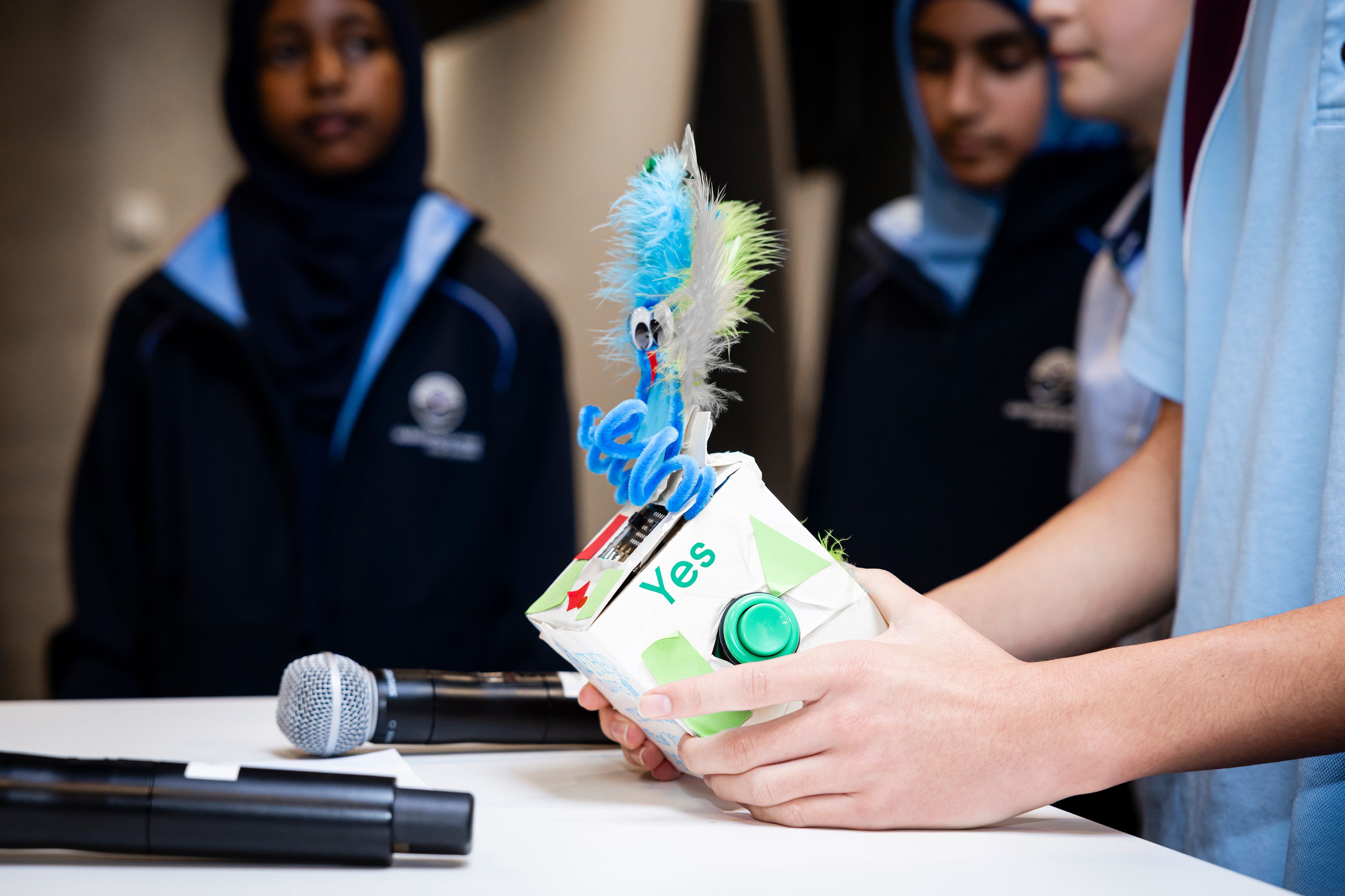 Close-up photo of a student holding a hand crafted cardboard device on a white table with 2 microphones. There are other students in the background.