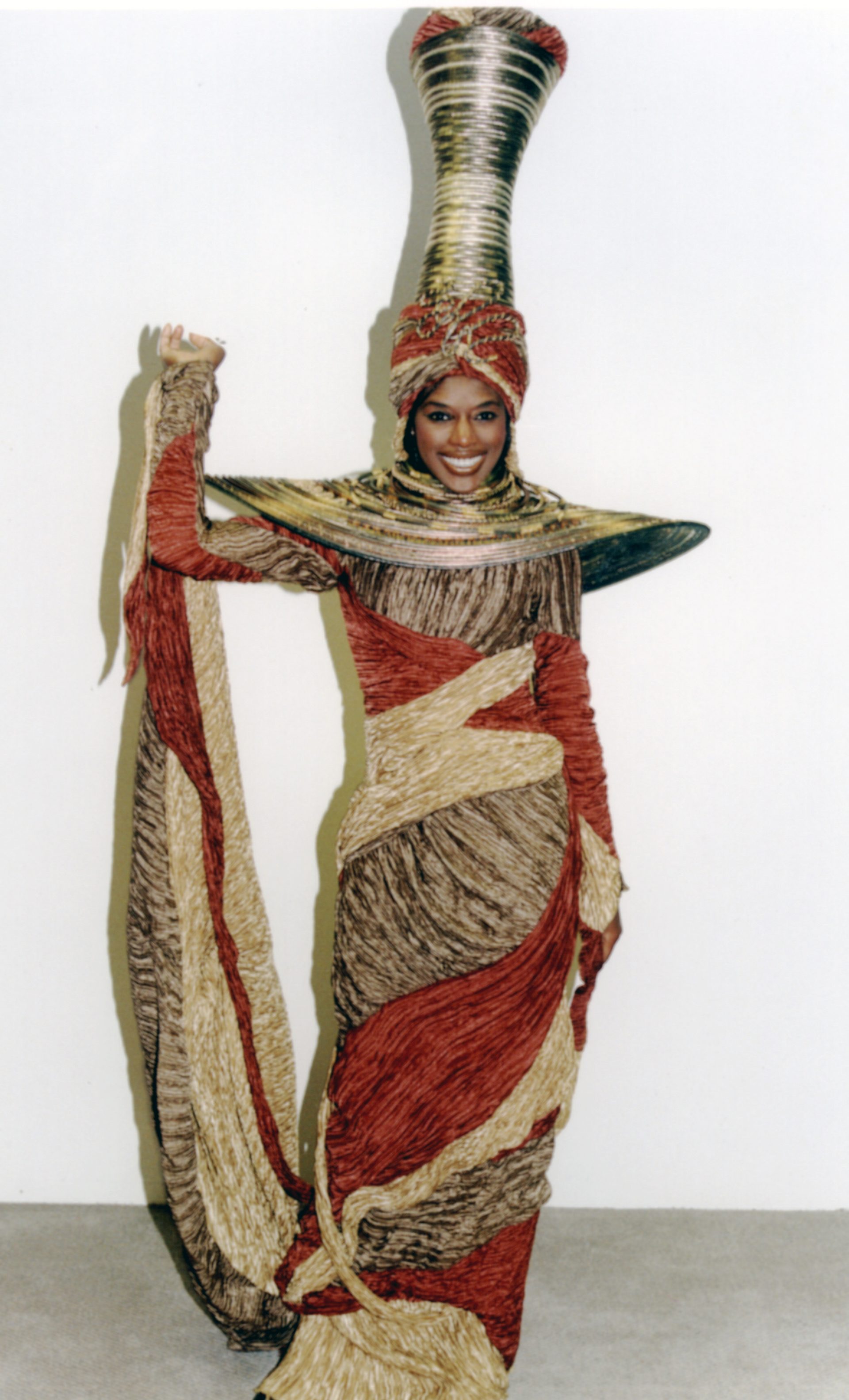 Woman wearing tall narrow gold hat with very wide collar is swathed in dress of red and gold crinkled material covering her feet and hold ups the train in her right hand.