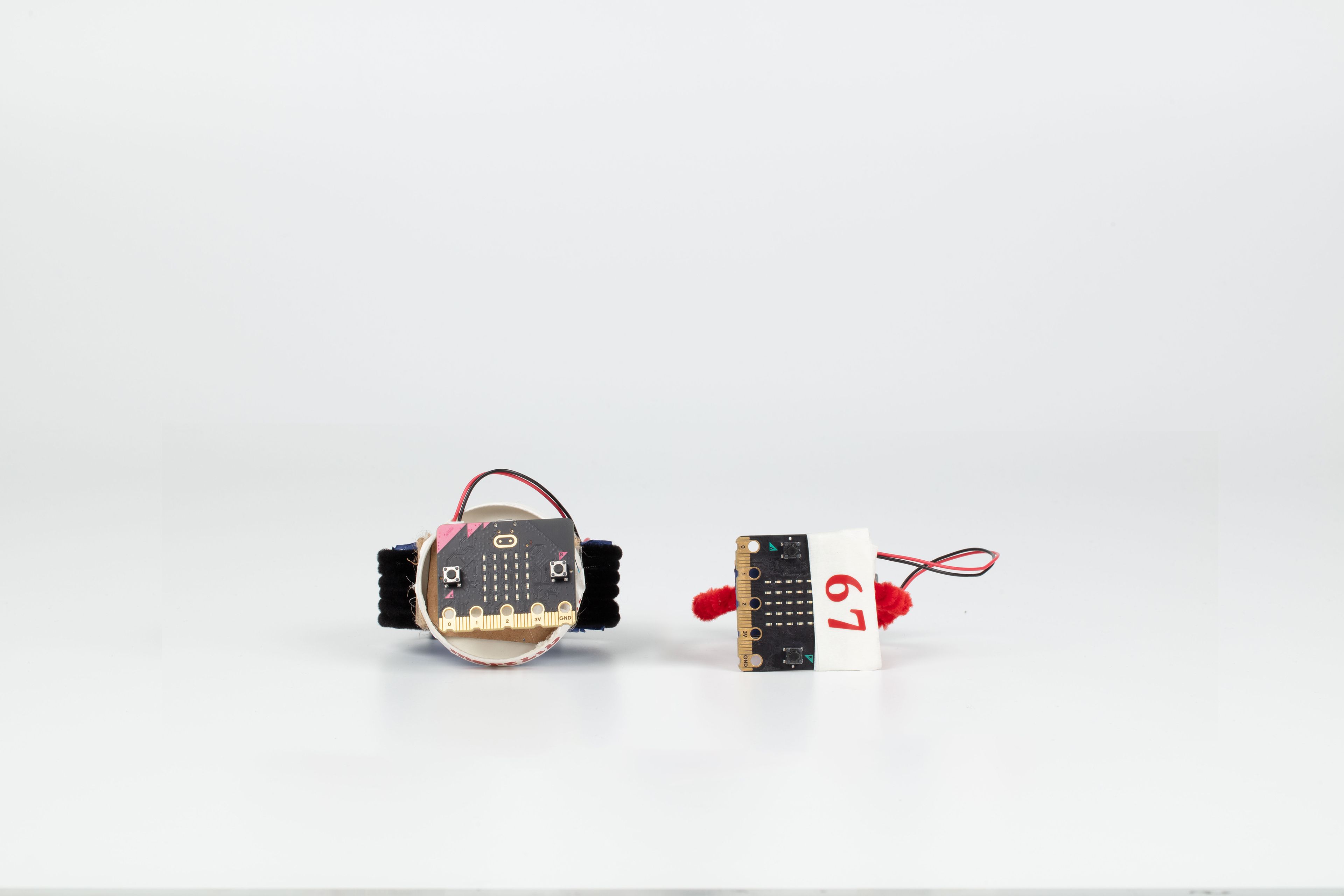 Image of a hand crafted mechanical device against a white background. Cardboard made watches using pipe cleaners as the wrist band, wires and small coding boards on the watchface.