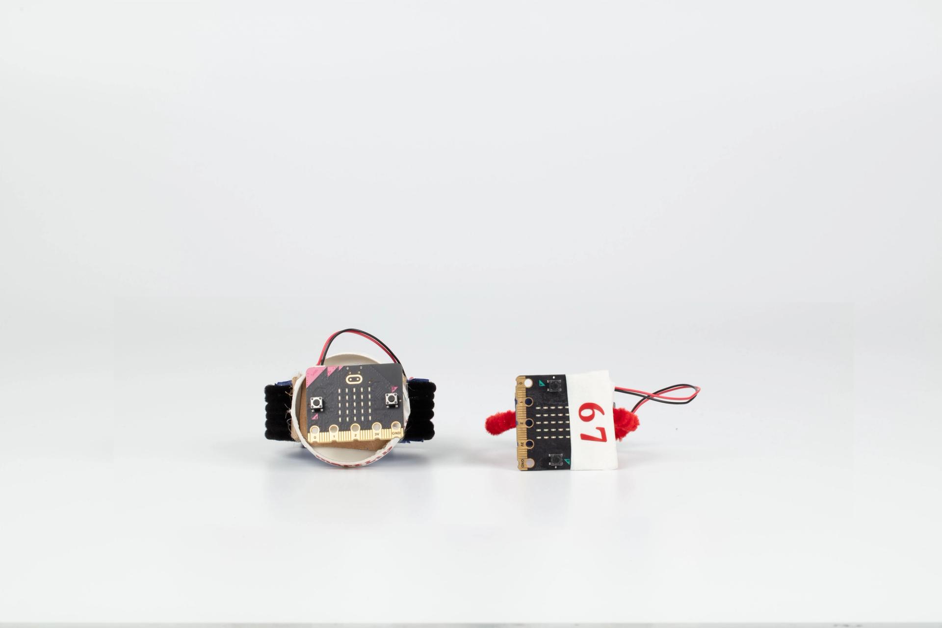 Image of a hand crafted mechanical device against a white background. Cardboard made watches using pipe cleaners as the wrist band, wires and small coding boards on the watchface.