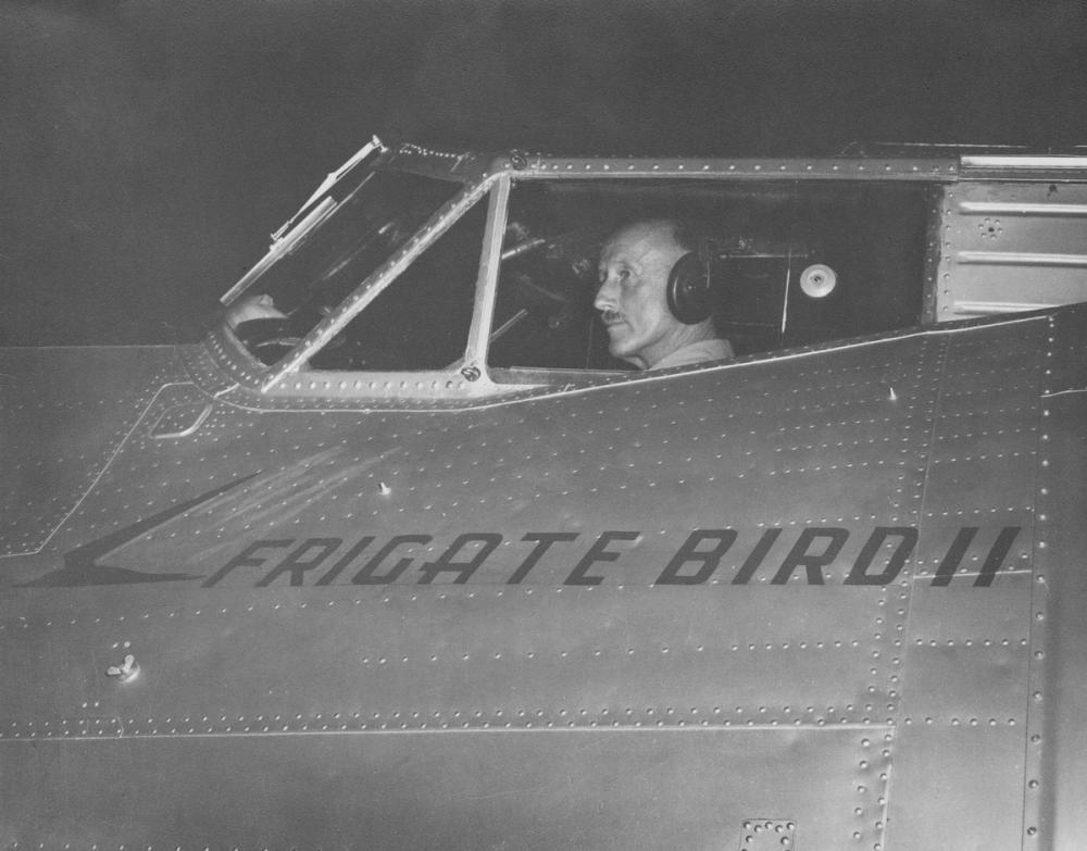 Pilot in cockpit of silver plane. The text Frigate Bird II is painted over metal rivets on the side of the aircraft.