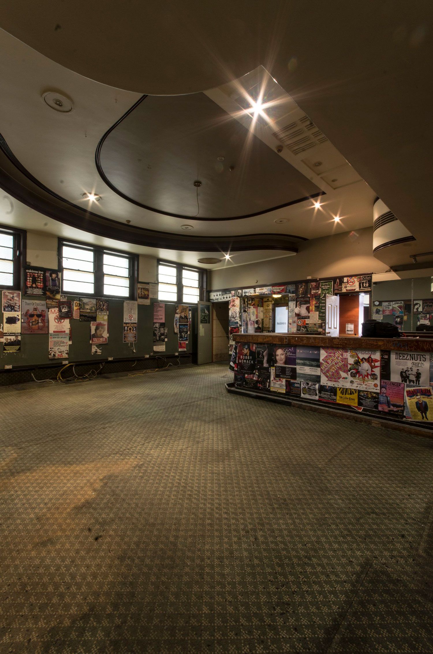 The interior of the old Clare Hotel. A big bar with a variety of posters on the wall. Some are falling down or damanged. The floor is a dirty green coloured carpet.