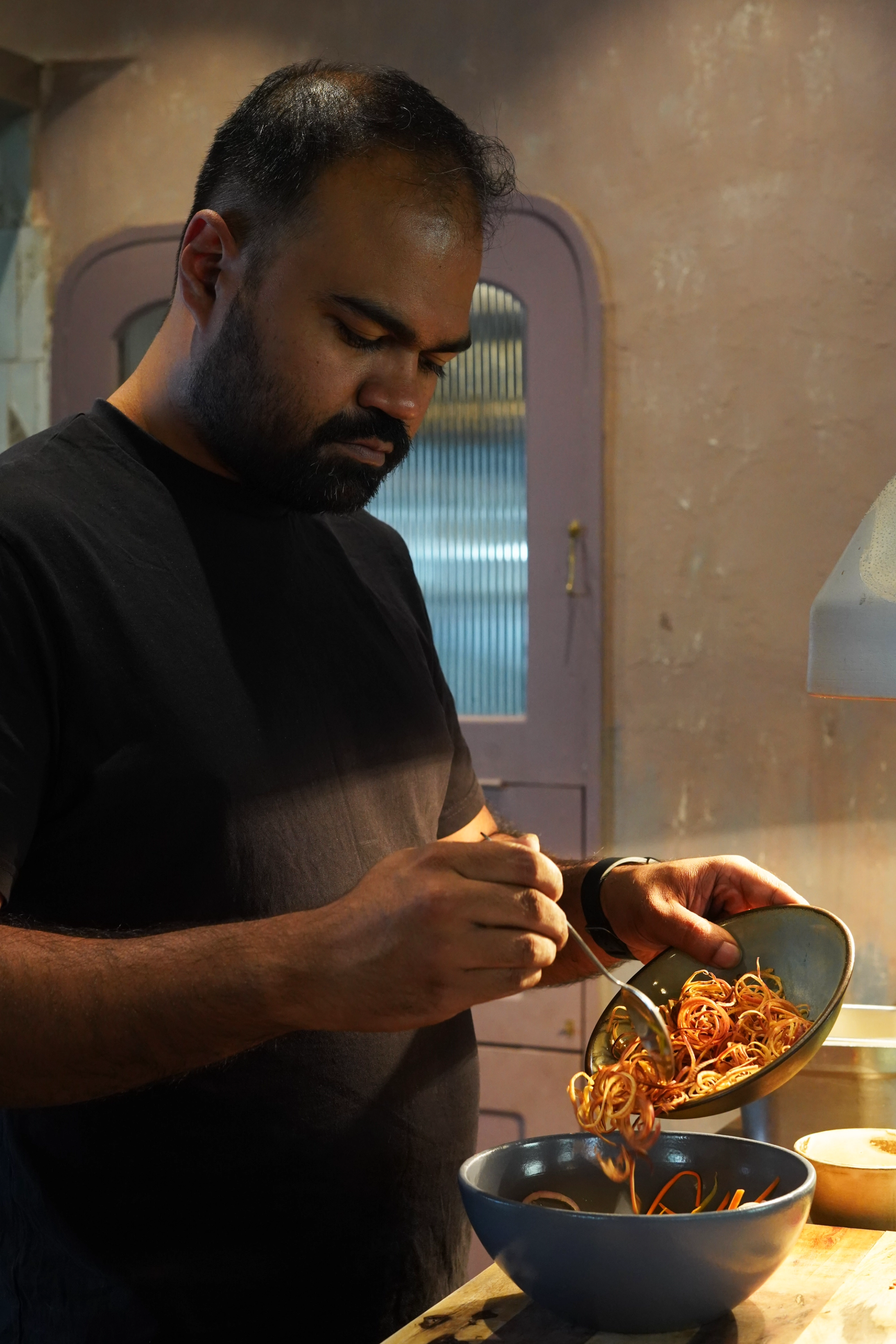 A Bengali chef transfers ingredients to a serving bowl. He wears a black T-shirt.