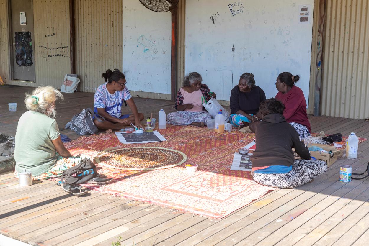 Six women sit outside, around a large woven mat.