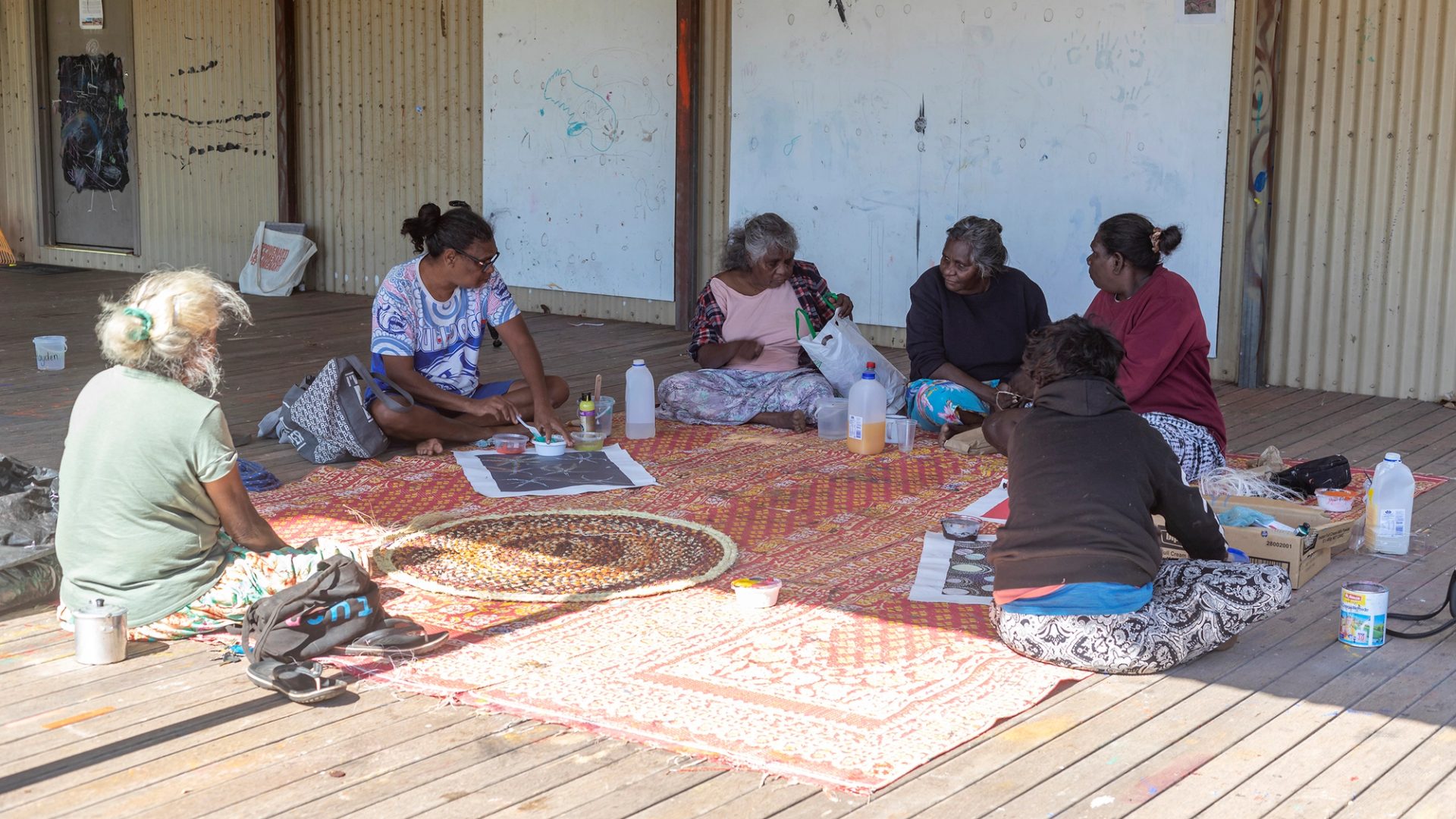 Six women sit outside, around a large woven mat.
