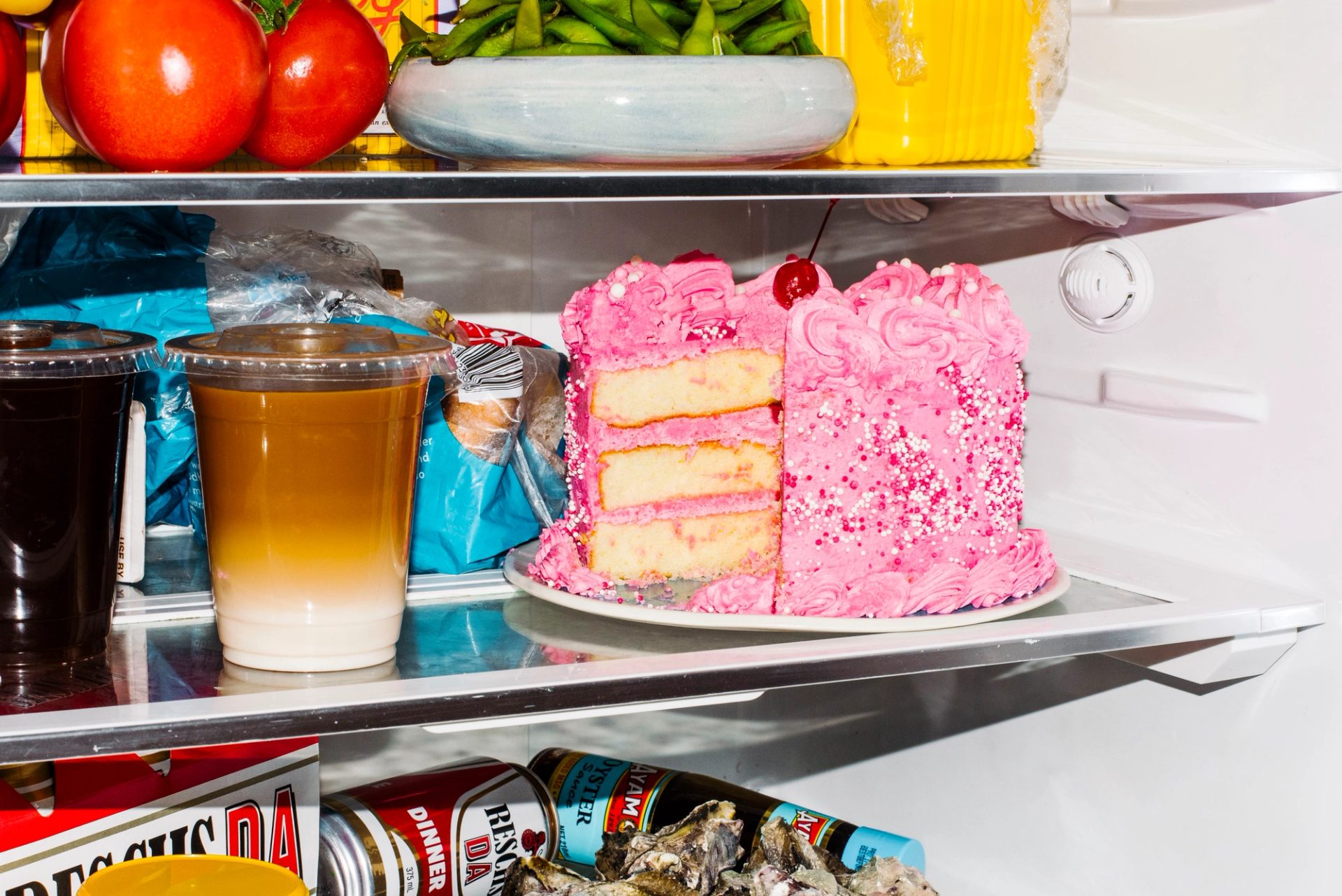 The middle shelves of an open fridge. On the lower shelf is a plate of oysters, a red can of Resch’s beer, two yellow Bonsoy long-life milk cartons, turned on their side. In front of them is a iced latte, in a clear plastic takeaway cup. On the shelf above are two more iced coffees, both in takeaway cups. To their left a red and green Fountain Tomato Sauce bottle lies on its sides. Slightly behind them and to the right is a carton of Oak Iced Coffee. Sitting on top of that is an almost empty bread bag. In front of the bread bag is a round sponge cake with pink icing and a cherry on top. A large slice has been cut from the right side of the cake.