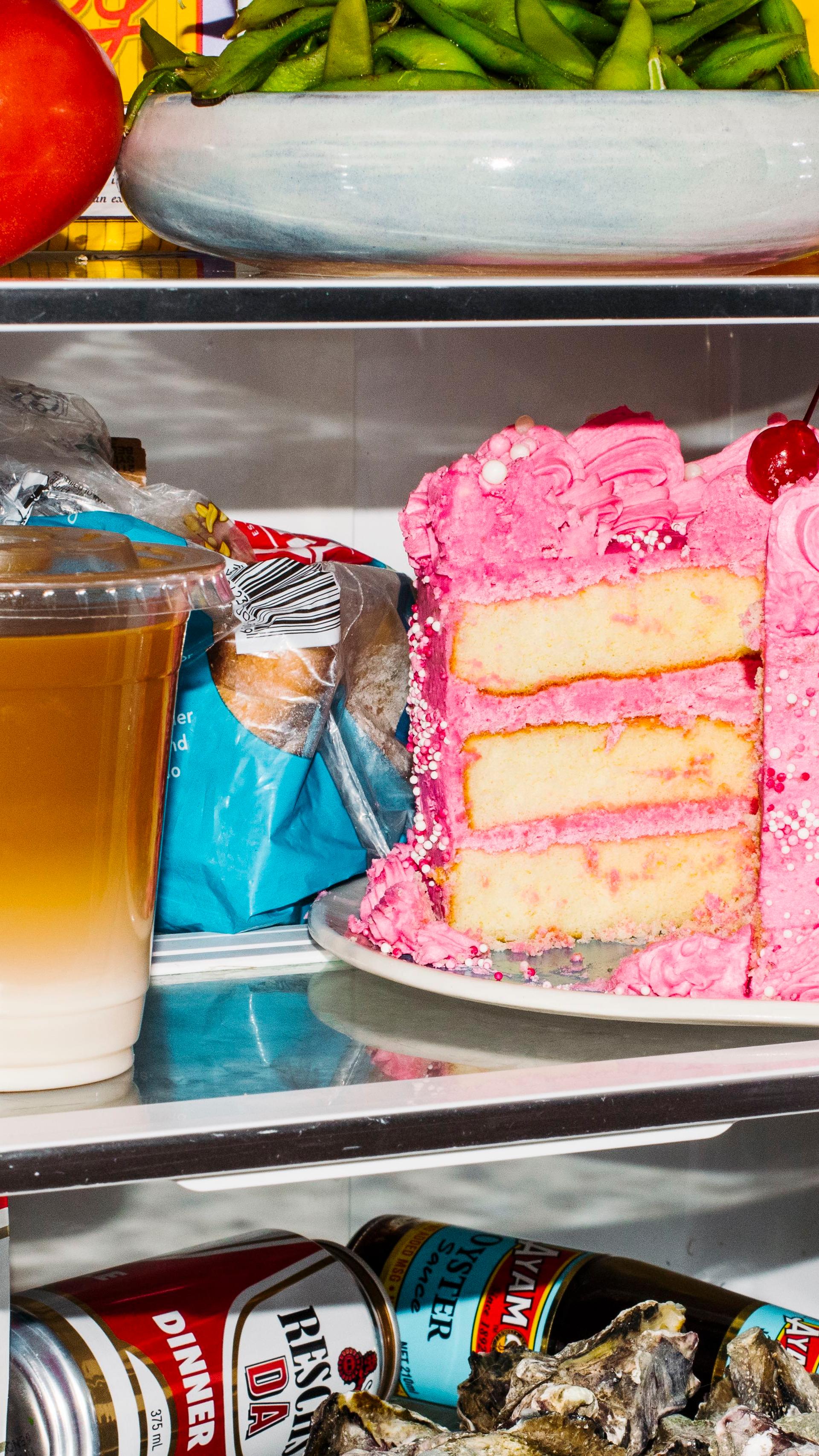 The middle shelves of an open fridge. On the lower shelf is a plate of oysters, a red can of Resch’s beer, two yellow Bonsoy long-life milk cartons, turned on their side. In front of them is a iced latte, in a clear plastic takeaway cup. On the shelf above are two more iced coffees, both in takeaway cups. To their left a red and green Fountain Tomato Sauce bottle lies on its sides. Slightly behind them and to the right is a carton of Oak Iced Coffee. Sitting on top of that is an almost empty bread bag. In front of the bread bag is a round sponge cake with pink icing and a cherry on top. A large slice has been cut from the right side of the cake.