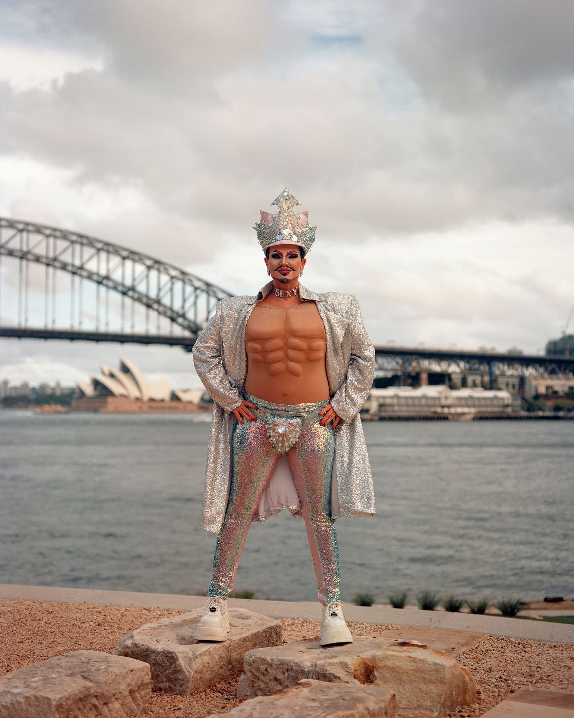 A drag king wearing a sparkly crown, coat and leggings stands on two rocks in front of the water. They’re wearing a padded chest piece with pronounced pectoral and abdominal muscles. The Sydney Harbour Bridge is visible in the background.