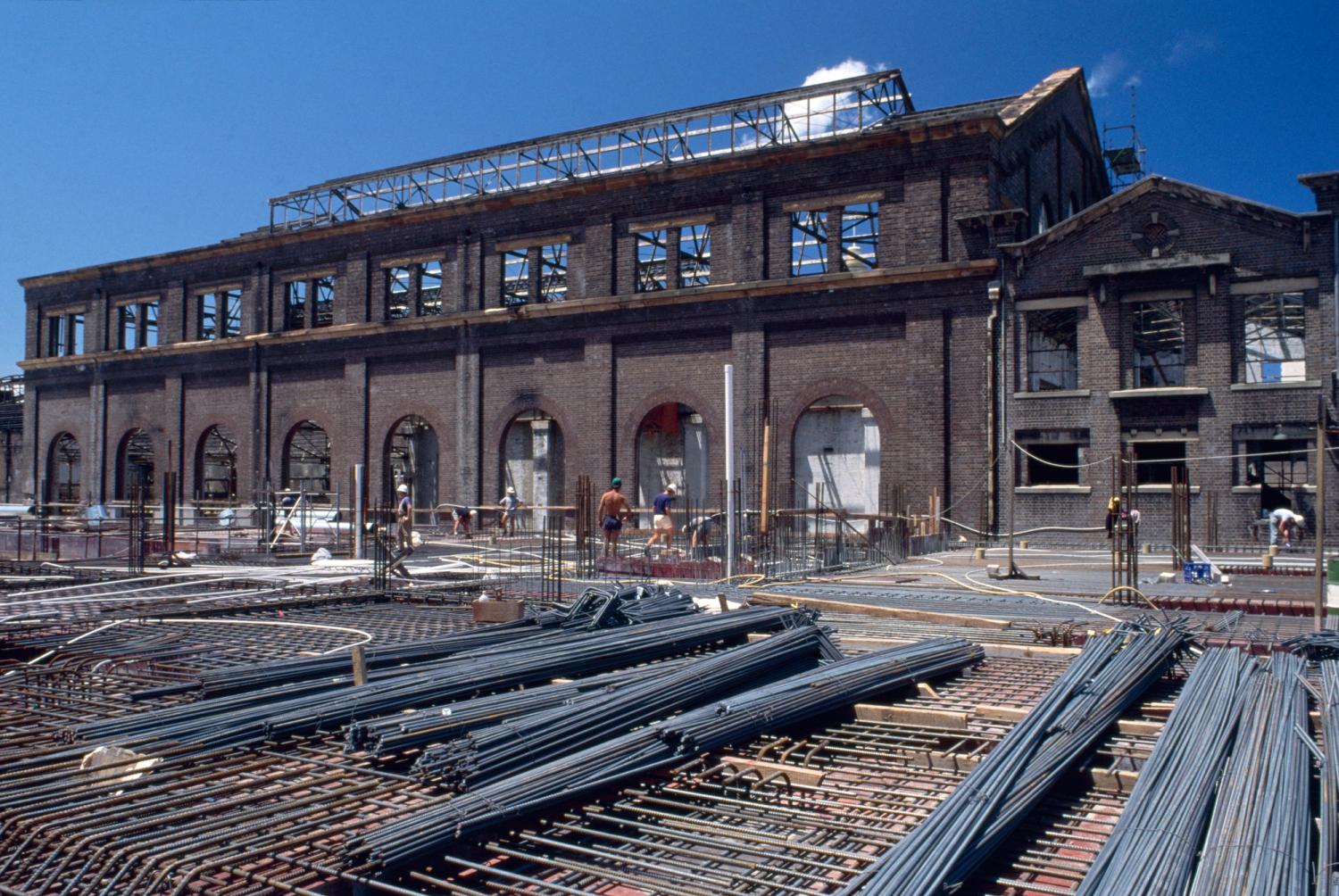 Colour Photograph of construction site with steel bars in the foreground, and a gutted brick building in the background and construction works walking through the frame