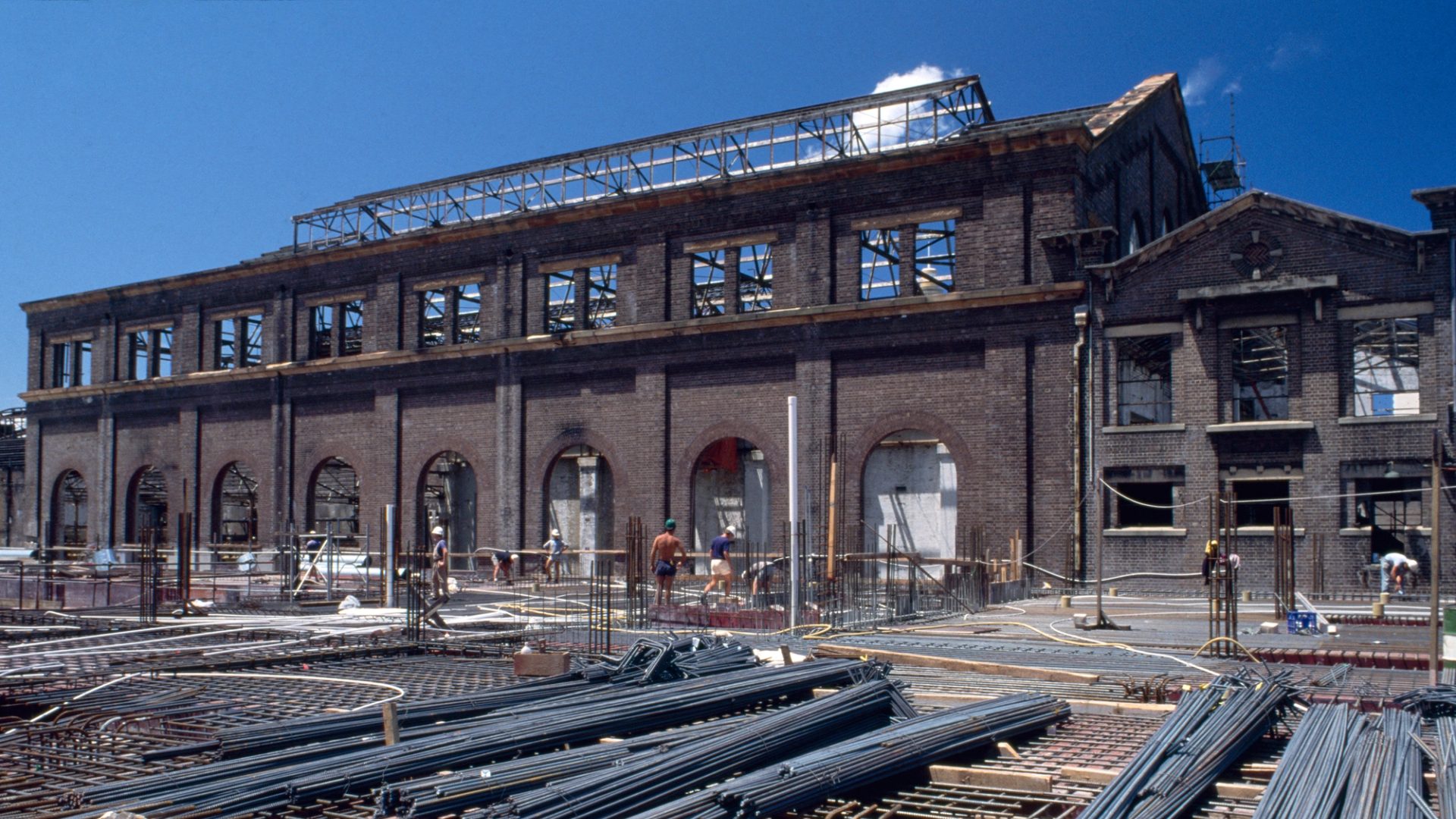 Colour Photograph of construction site with steel bars in the foreground, and a gutted brick building in the background and construction works walking through the frame