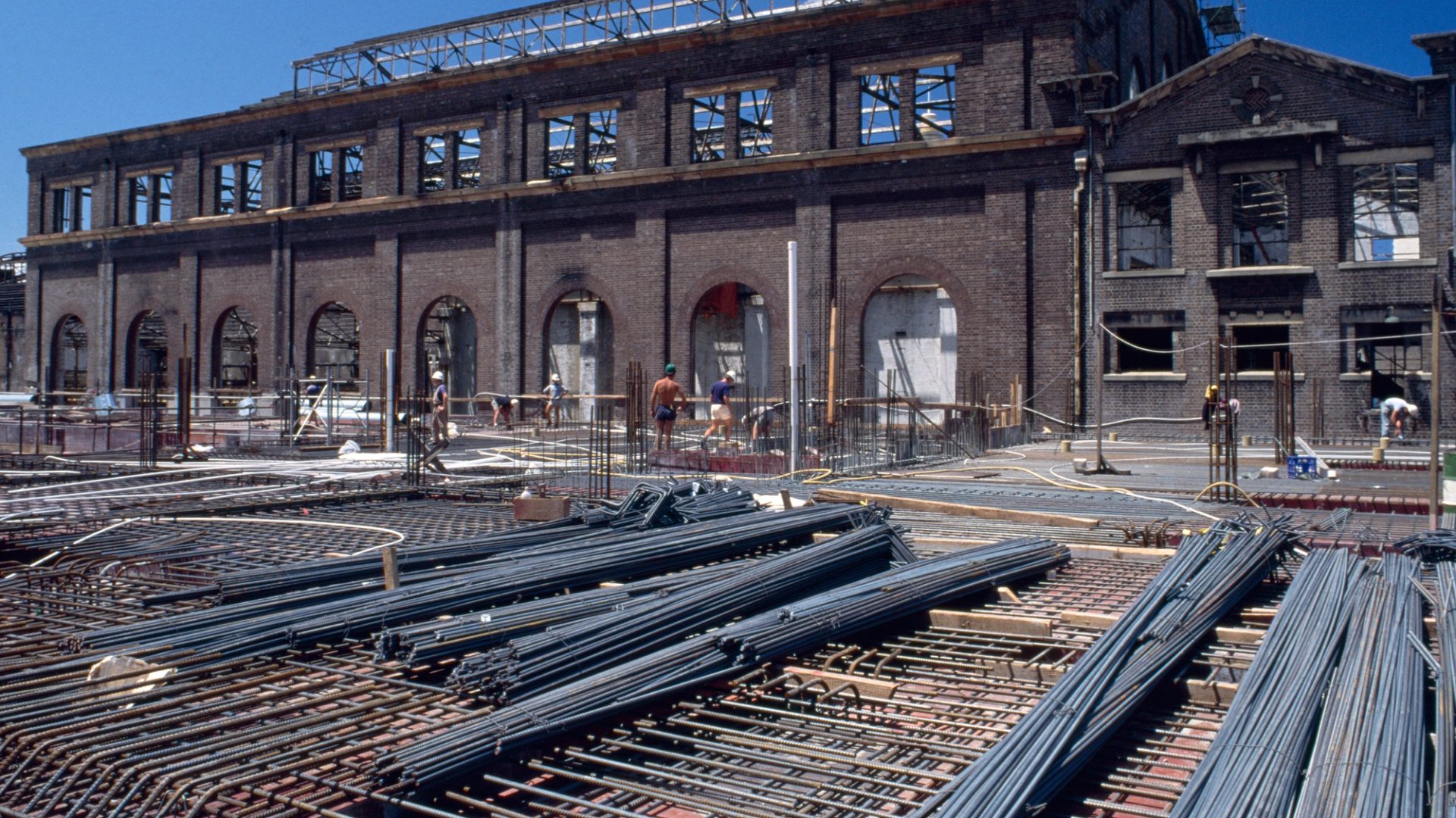 Colour Photograph of construction site with steel bars in the foreground, and a gutted brick building in the background and construction works walking through the frame