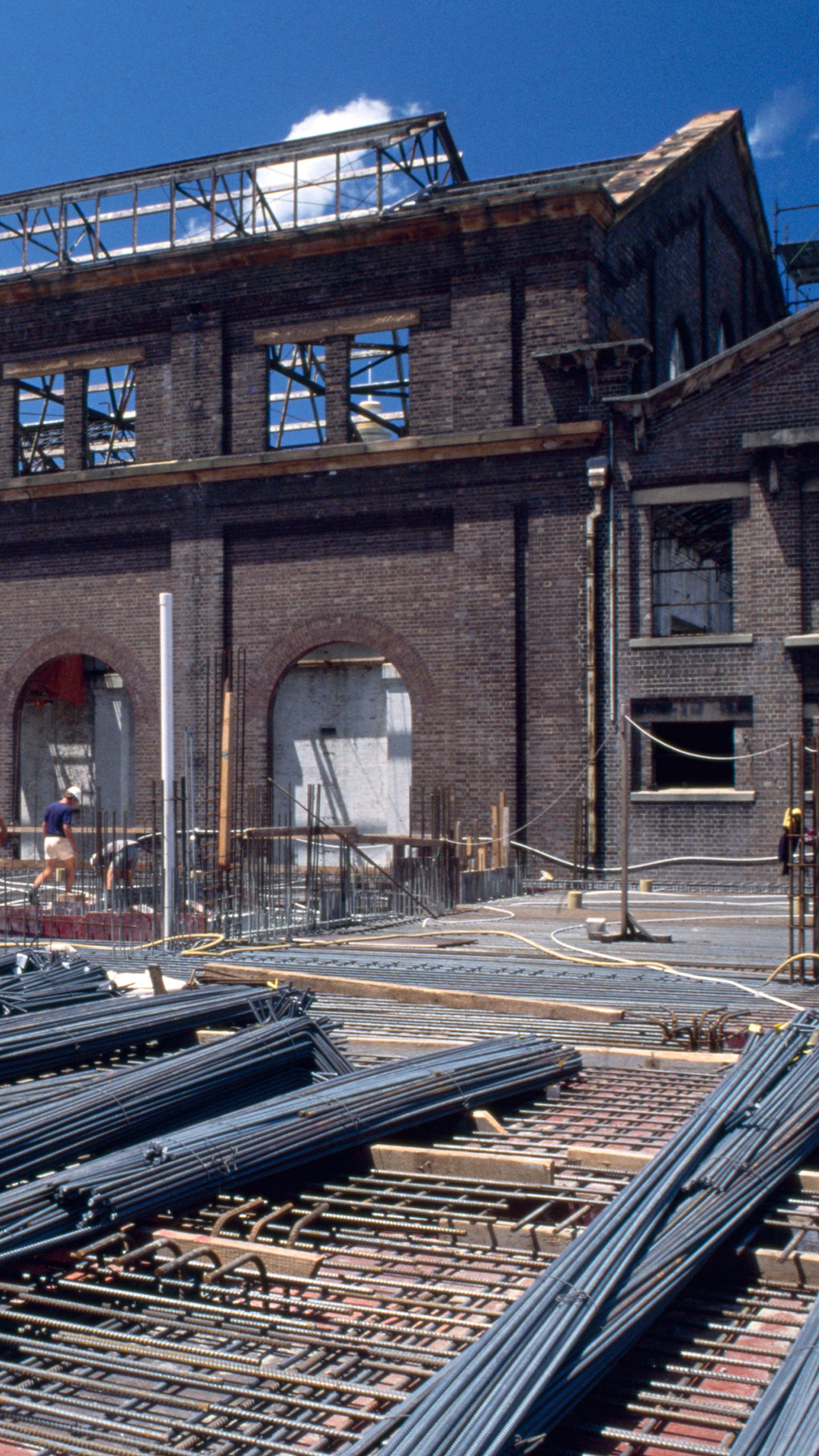Colour Photograph of construction site with steel bars in the foreground, and a gutted brick building in the background and construction works walking through the frame