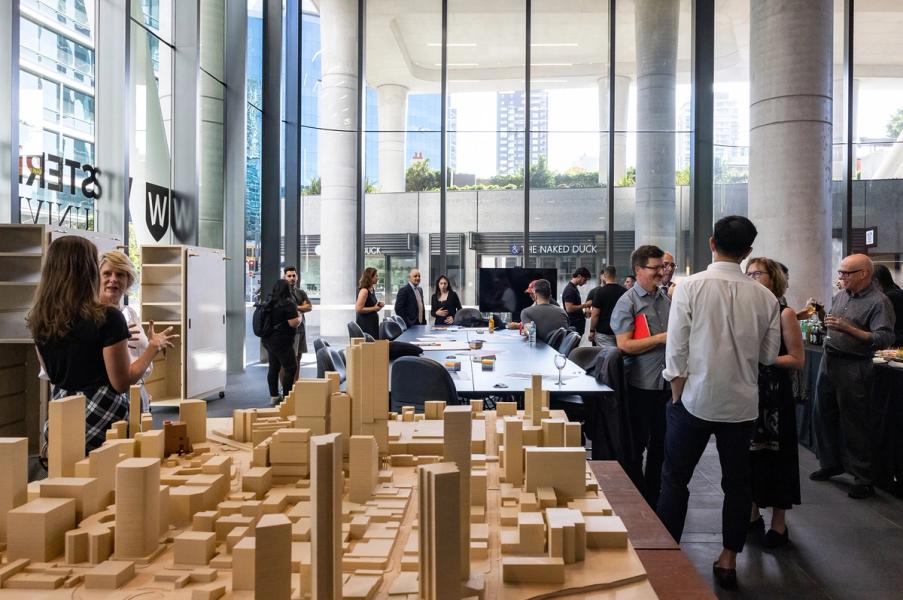 Wooden model of city in foreground while humans stand and gather around a large meeting desk in a room with floor to ceilings windows.