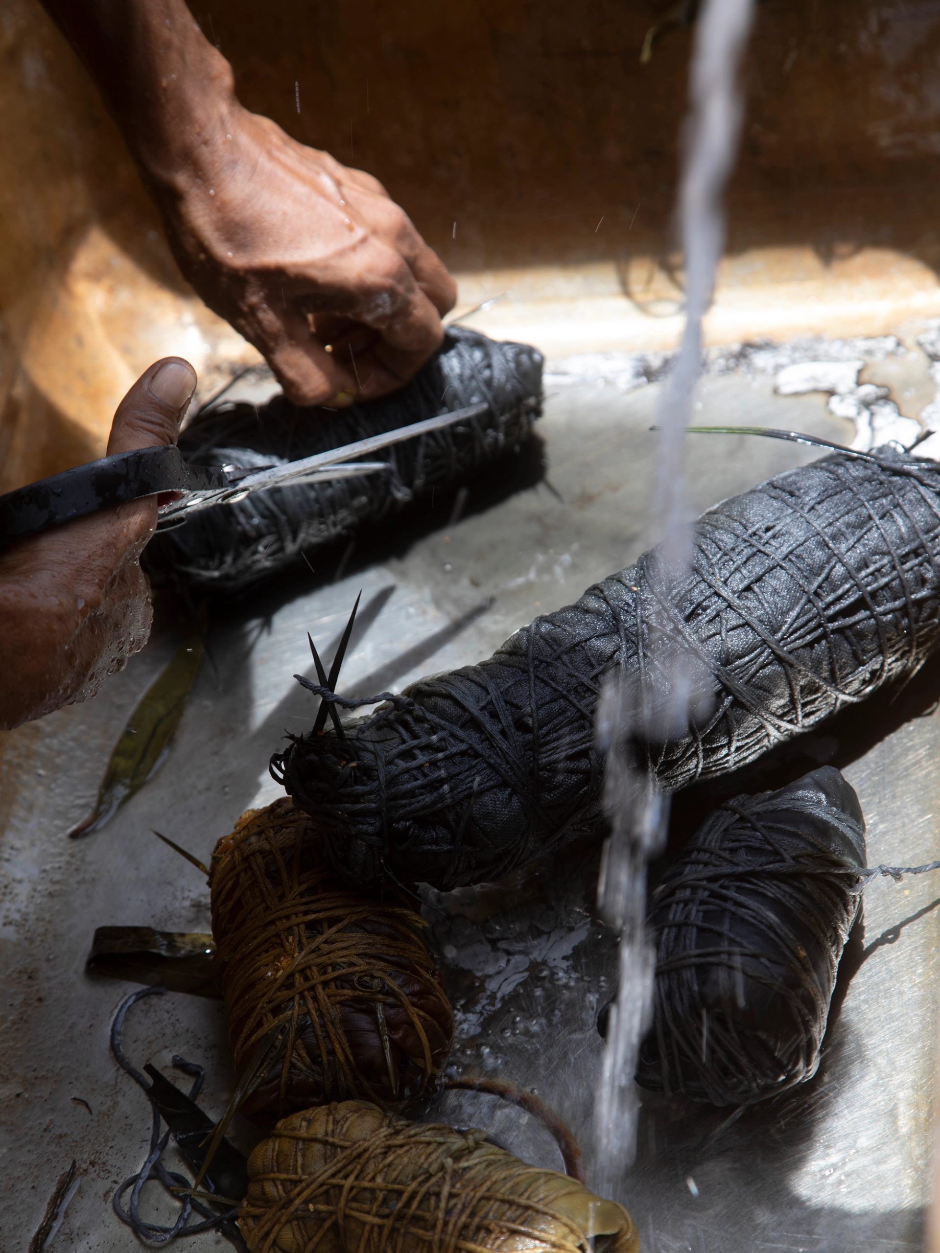 Five black and brown cylindrical fabric bundles wrapped in string. Two hands at the top left of the frame are holding a pair of scissors.