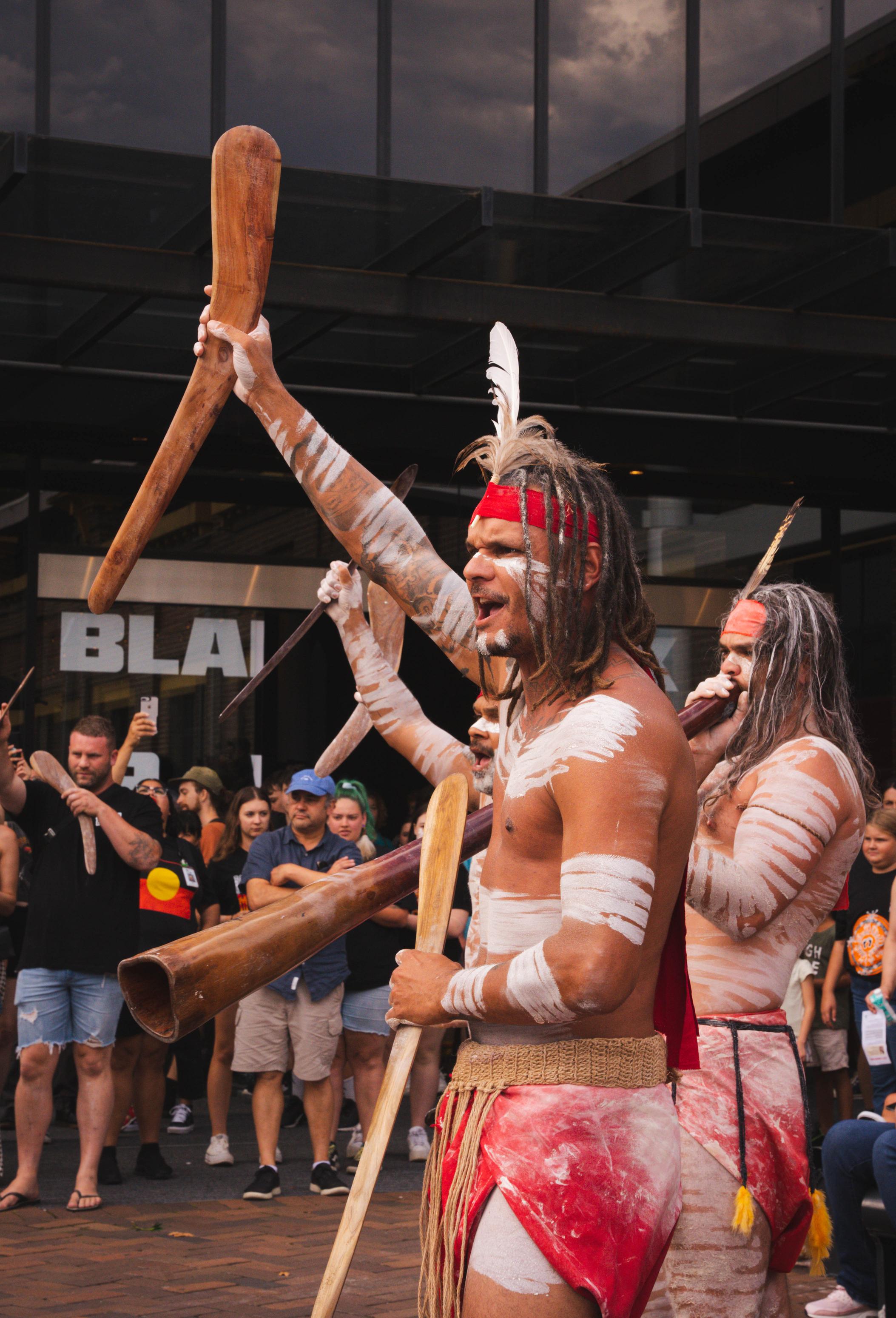 A First Nations performer in traditional dress is mid performance and is holding boomerang in his right hand.