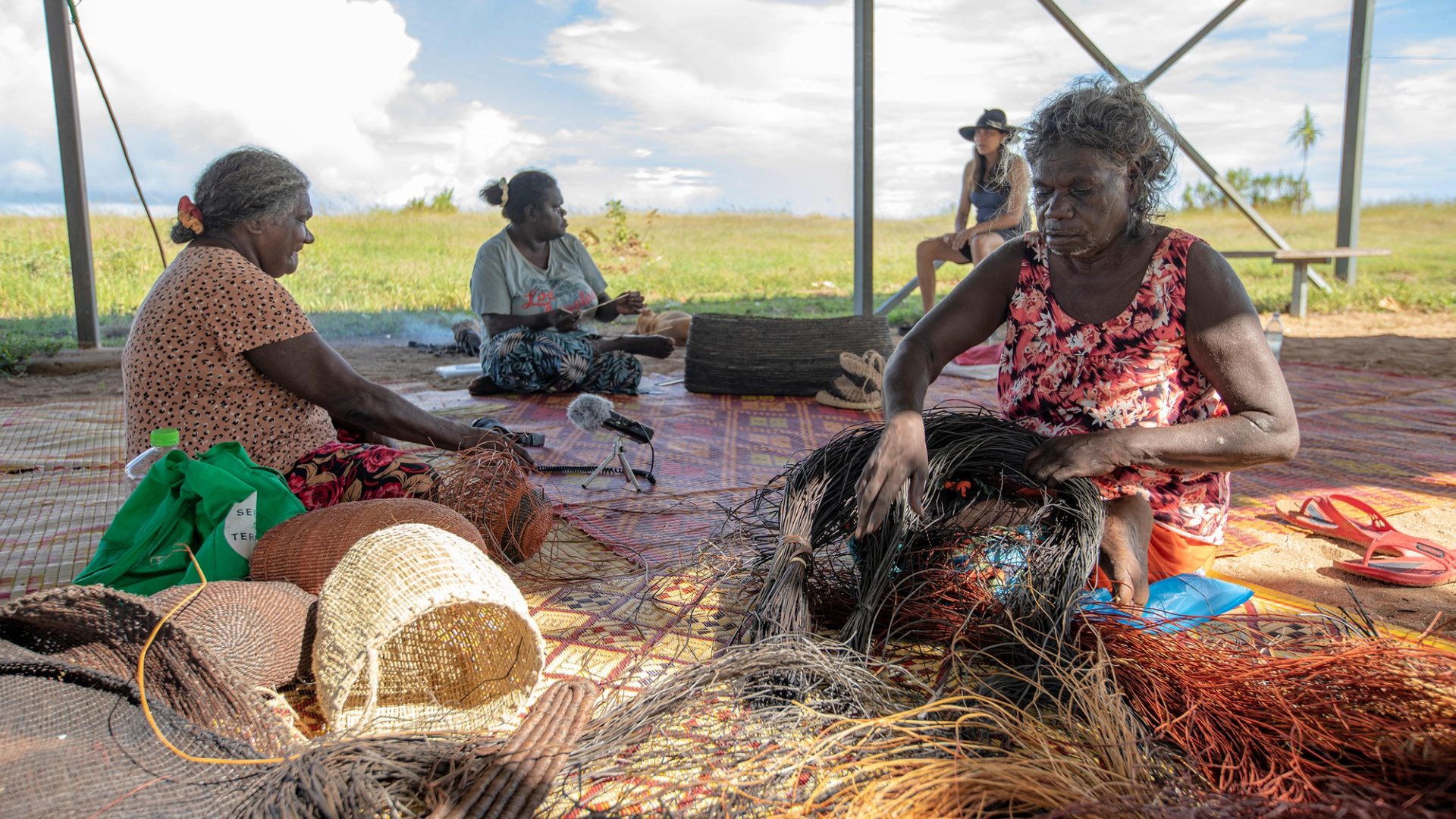Four women sit at different spots outside, on a large woven mat, weaving baskets