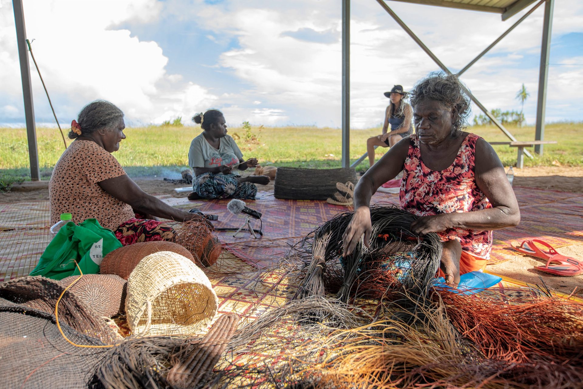 Four women sit at different spots outside, on a large woven mat, weaving baskets