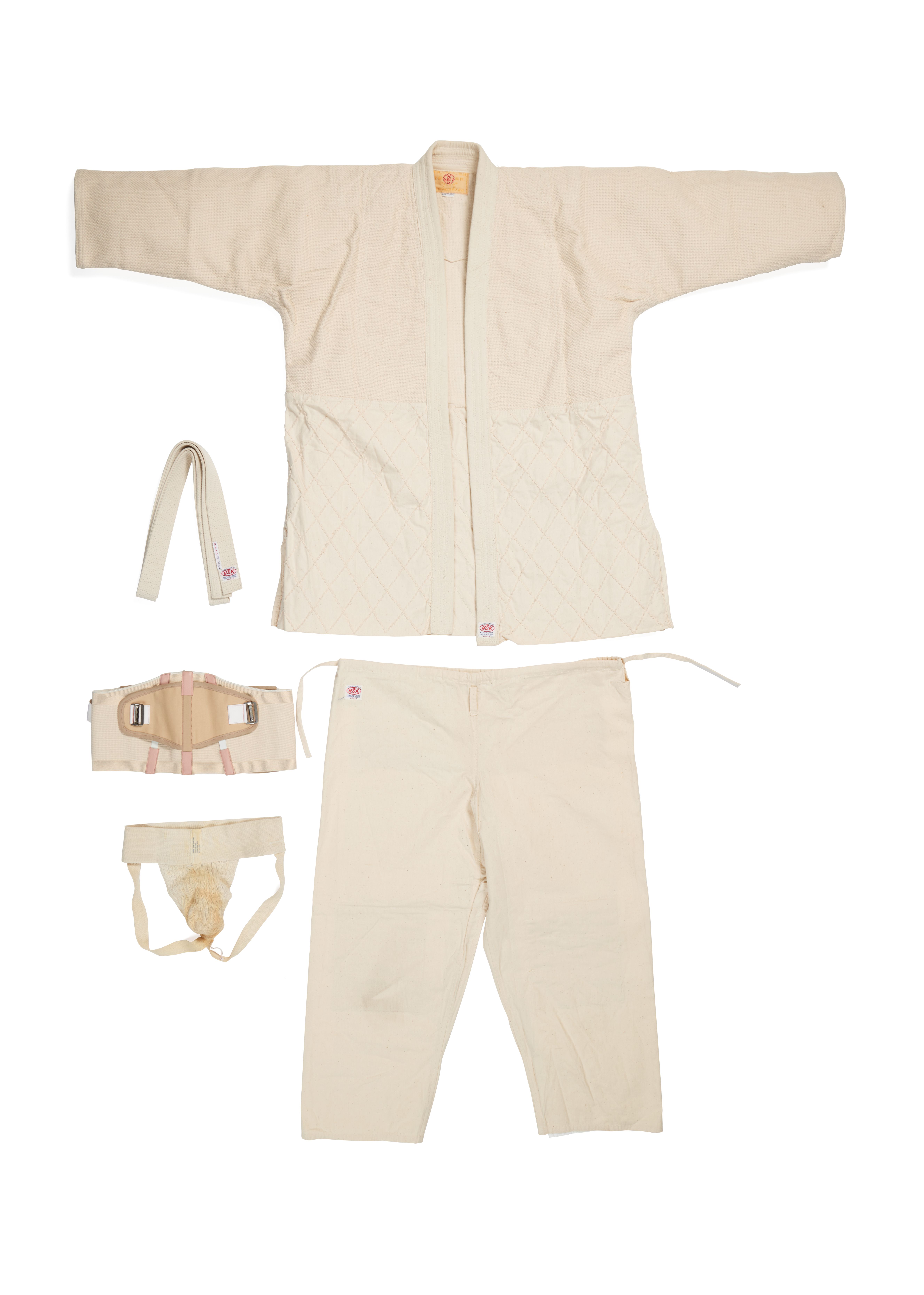 Photograph of a man's martial arts outfit made of cotton. A white wrap-over long sleeve shirt and loose pants matching fabrics. On the left are other accessories to the uniform such as a band and elastic accessories.