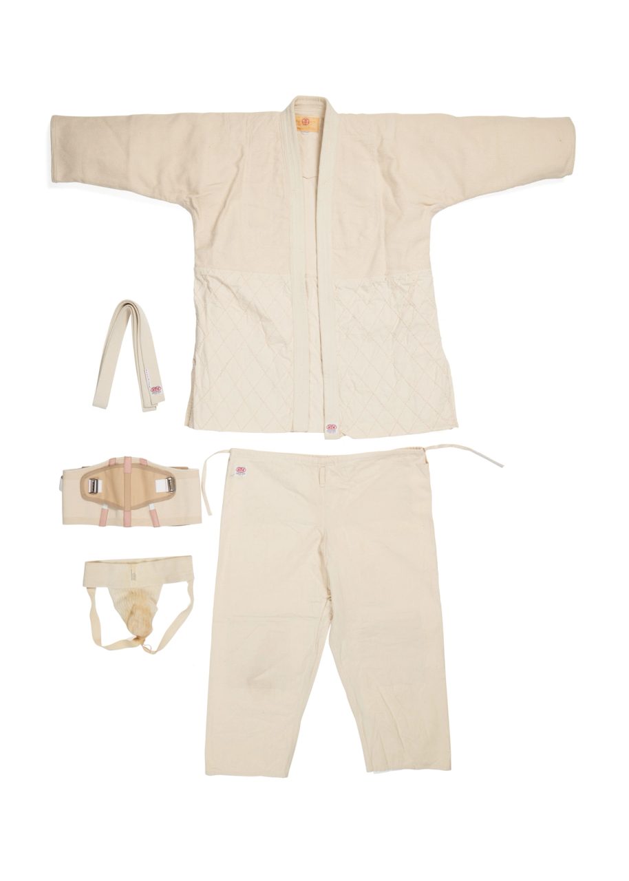 Photograph of a man's martial arts outfit made of cotton. A white wrap-over long sleeve shirt and loose pants matching fabrics. On the left are other accessories to the uniform such as a band and elastic accessories.