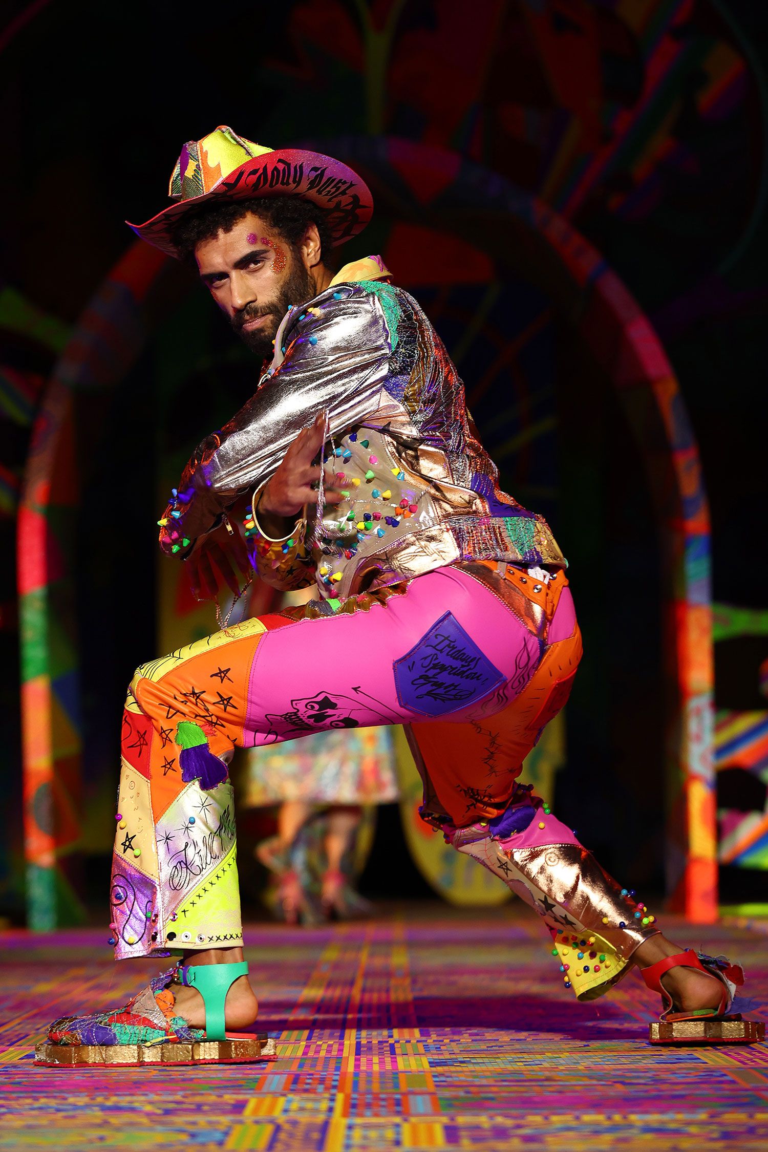 A model poses on the runway wearing a multicoloured outfit with a cowboy hat.