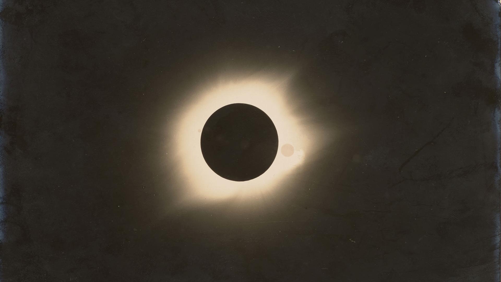 Black and white photograph of a total solar eclipse. Black circle in the centre with white glow around it, remainder of the image is black.