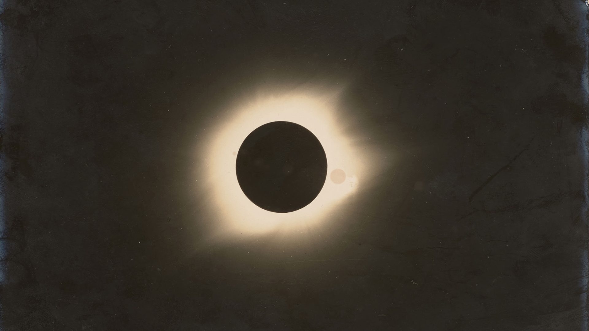 Black and white photograph of a total solar eclipse. Black circle in the centre with white glow around it, remainder of the image is black.