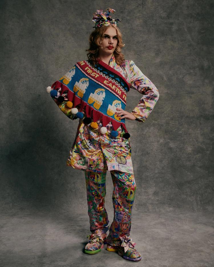 A model poses against a grey background wearing a multicoloured knitted top, patterned pants and a crown.