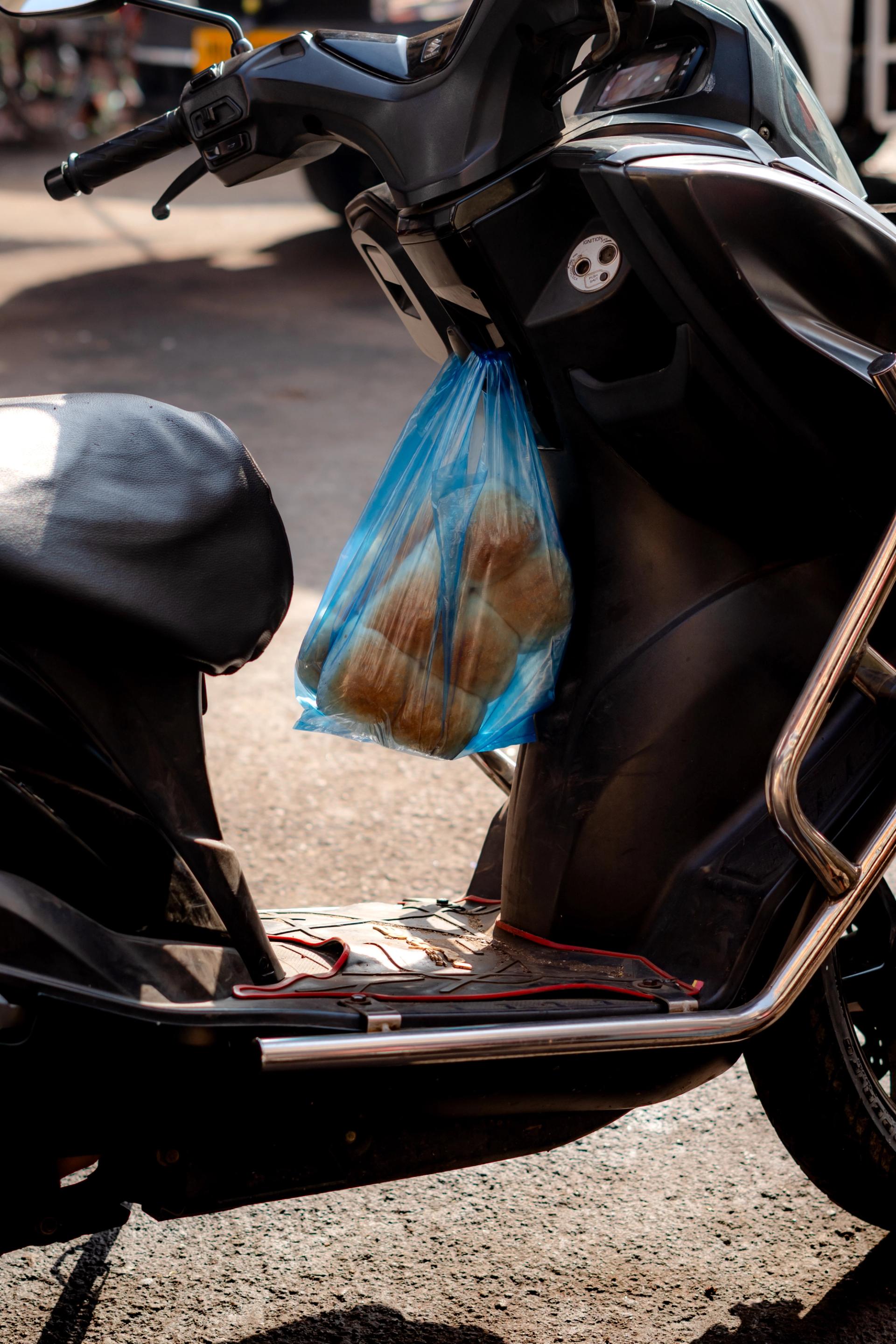 Two blue plastic bags, each with half a dozen bread rolls, hang on the inside hook of a Vespa.
