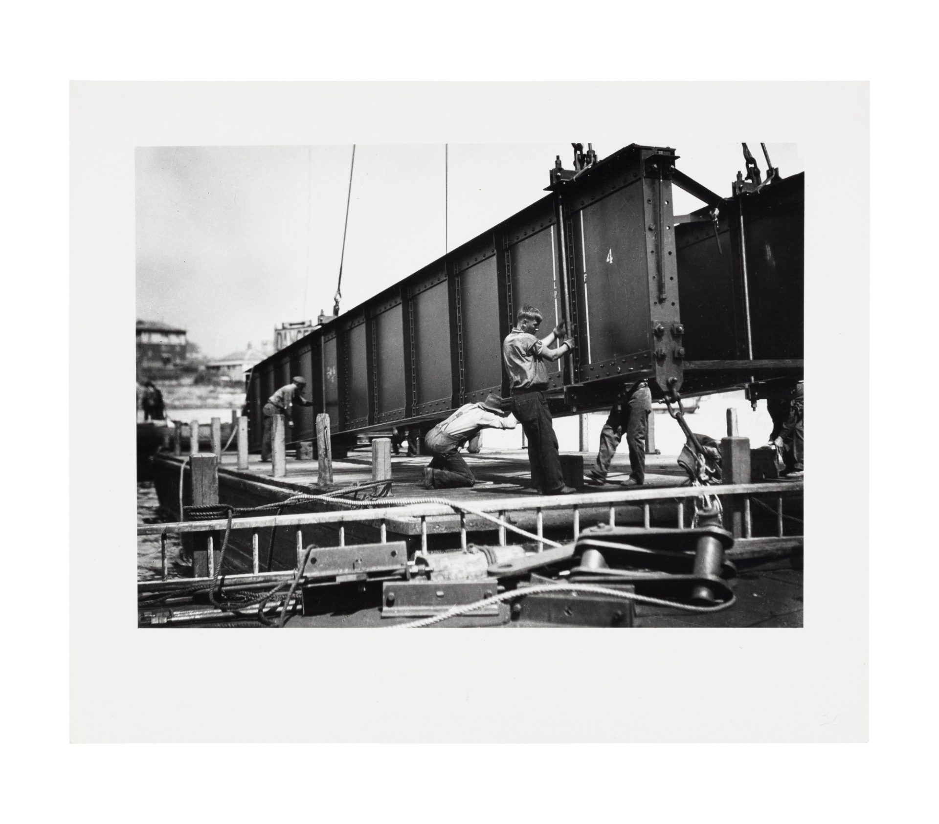 Image of four men supporting a bridge girder being lifted, most likely by a crane. They appear to be on a pier or barge on the water.