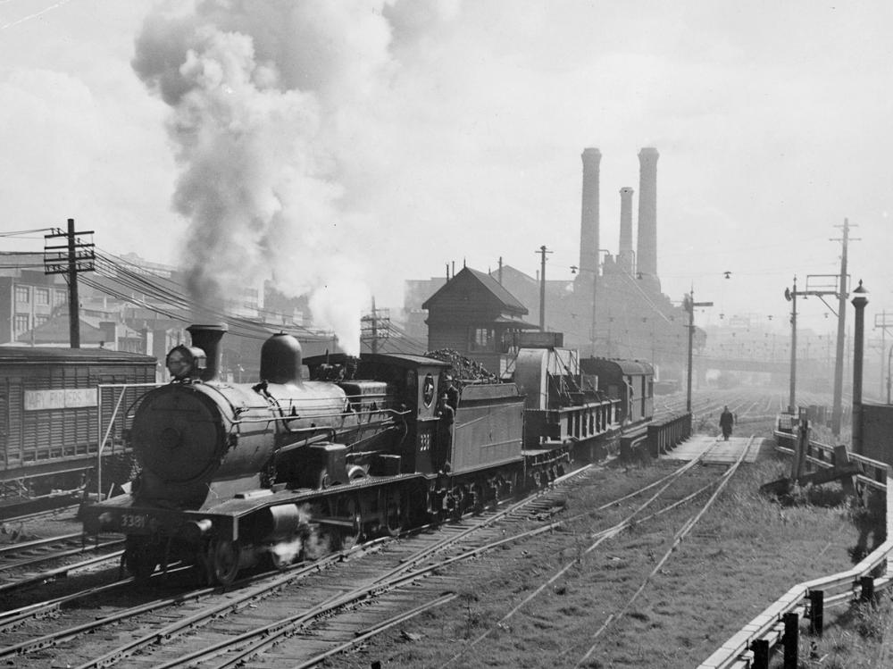 Black and white image of a steam train moving down a track with chimney stacks in background