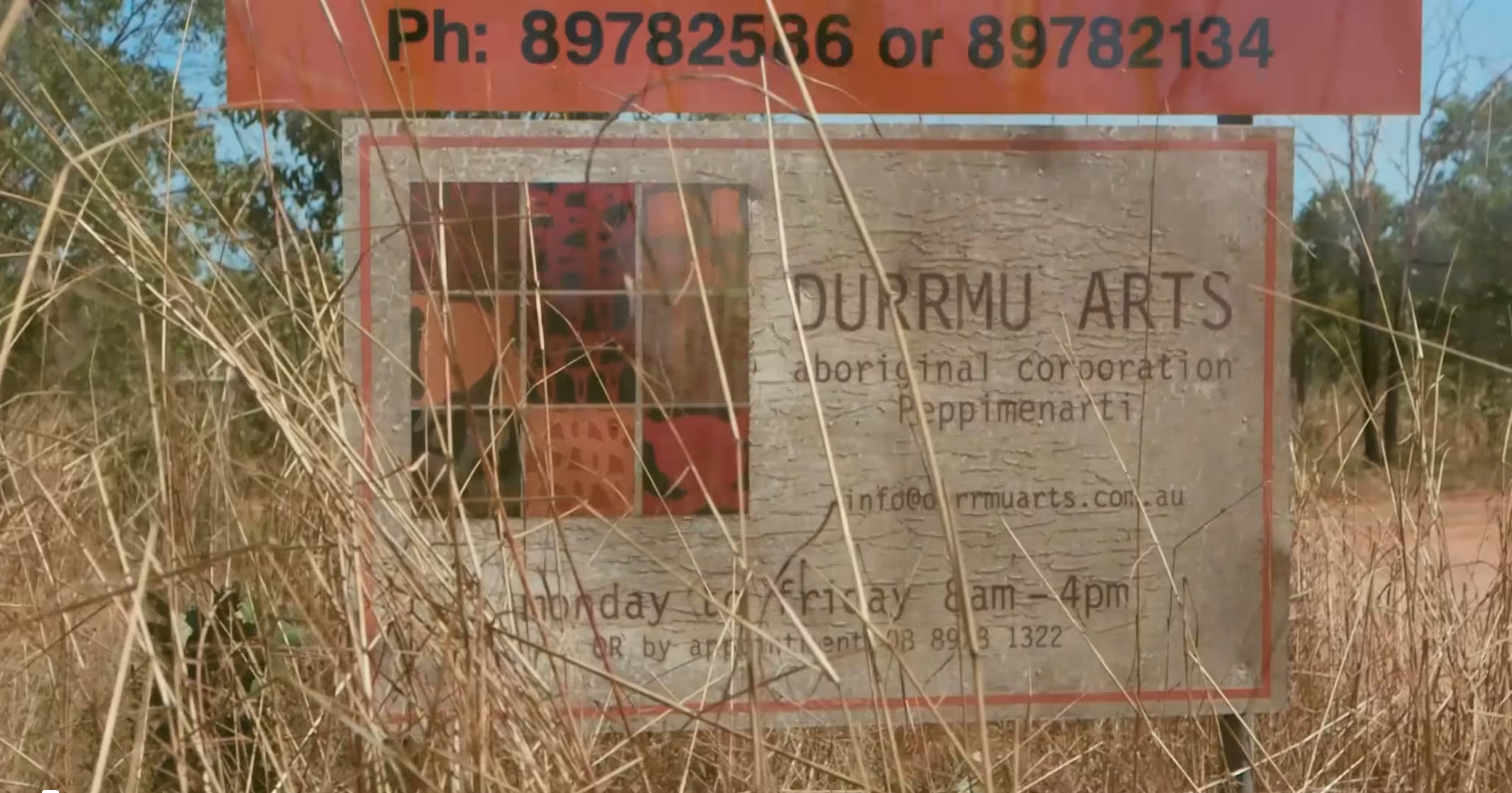 A worn rectangular sign behind long dry grass. There is a thin orange border around the sign edges. Text on the sign reads ‘DURRMU ARTS aboriginal corporation Peppimenarti info@durrmuarts.com.au monday to friday 8am-4pm OR by appointment 08 8918 1322’. On top of the sign is a longer, and thinner orange rectangle with thick black text reading ‘Ph: 89782586 or 89782134.’