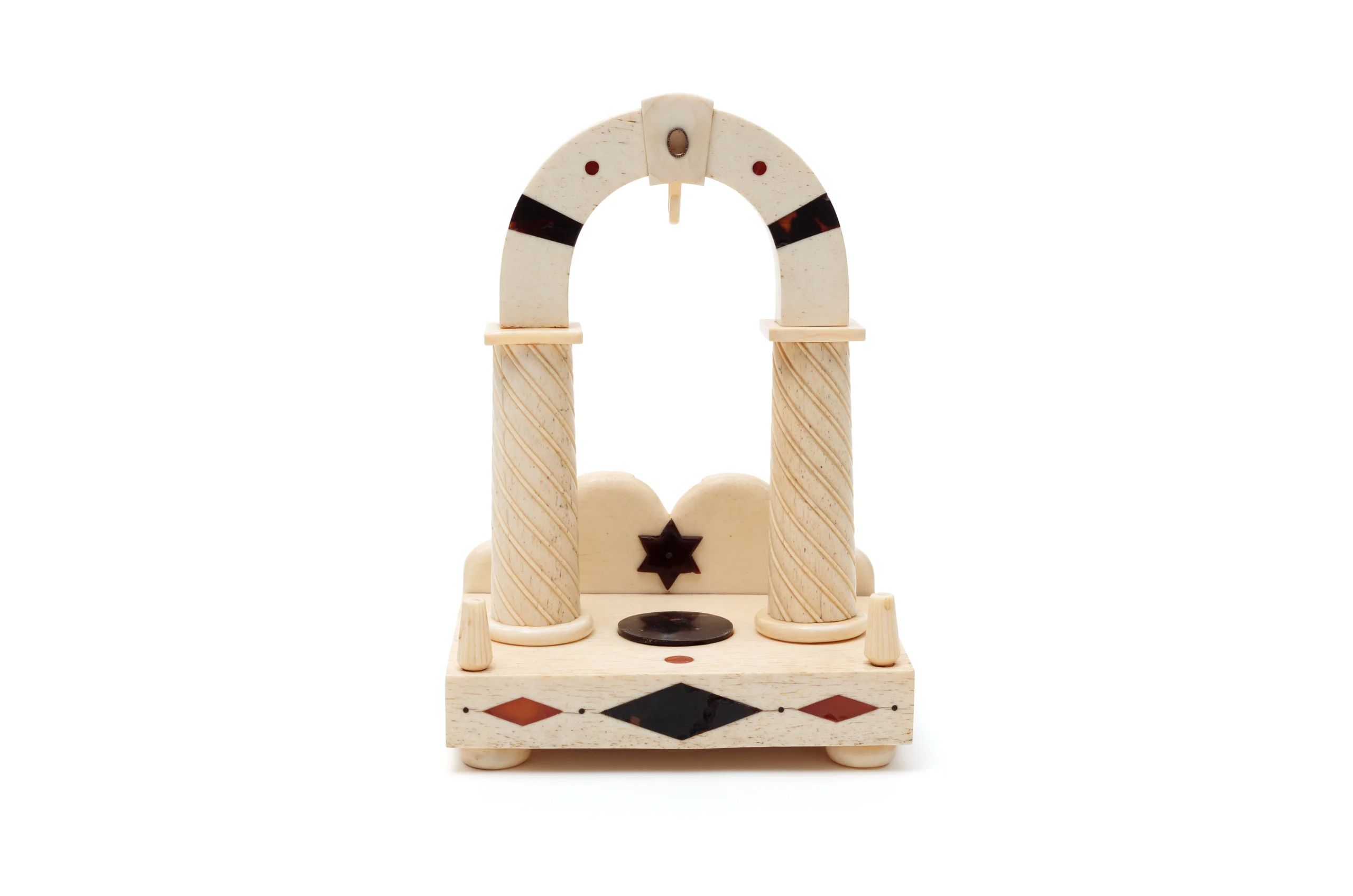 A pale bone-coloured arched object with two columns on a bone-coloured stand, all decorated with coloured inlay, and a backing board with an inlaid star.