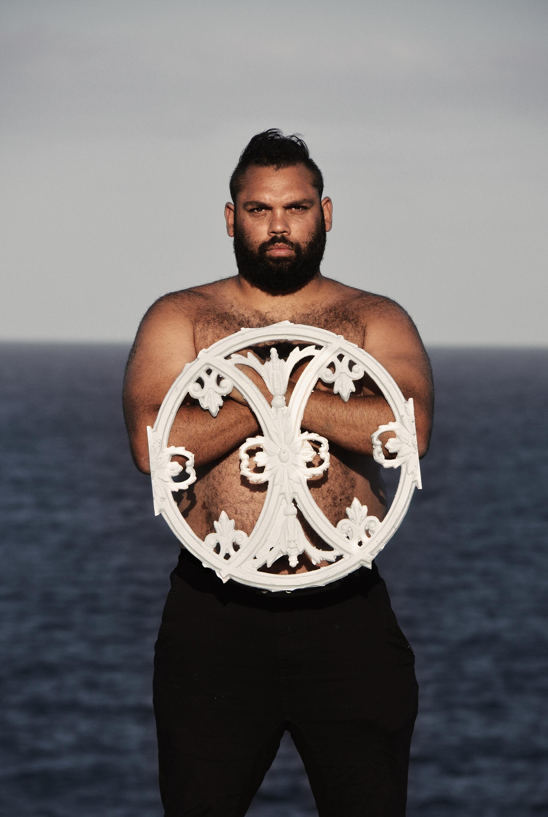 A shirtless man with dark short hair and a dark beard holds white circular fence heraldry.