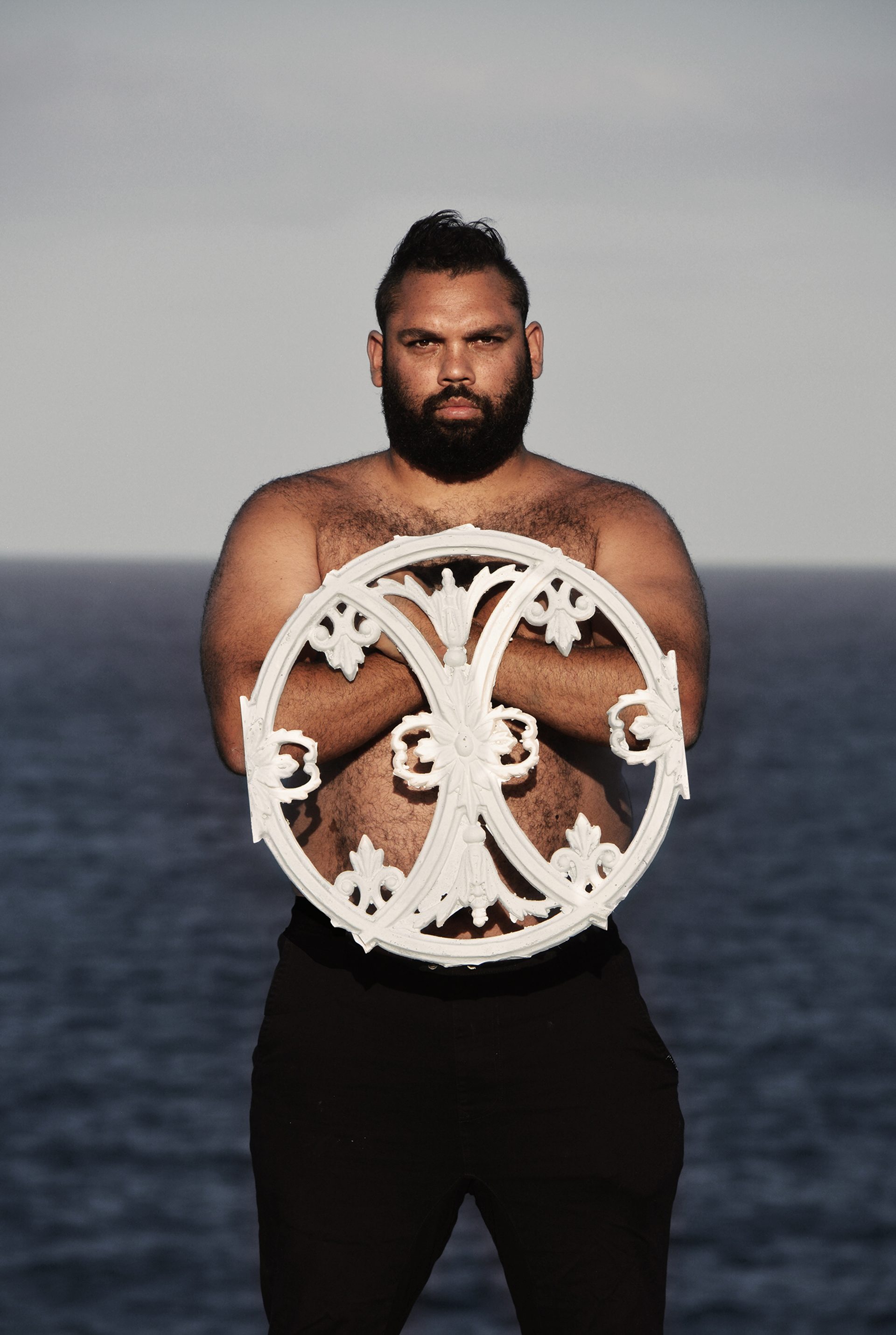 A shirtless man with dark short hair and a dark beard holds white circular fence heraldry.