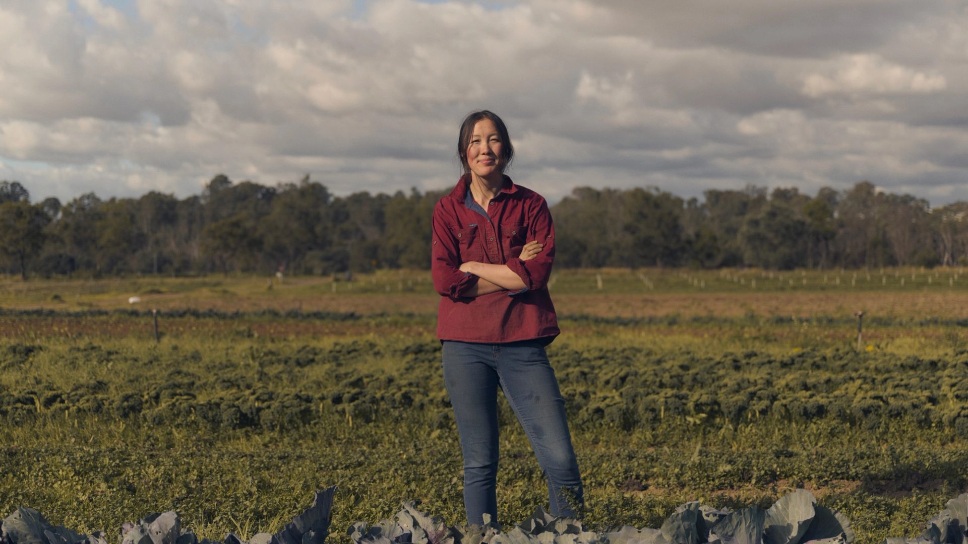 A woman stands confidently in a vibrant green field, surrounded by rows of leafy crops. She is wearing a red button-up shirt and jeans, with her arms crossed as she smiles at the camera. The background features expansive farmland under a partly cloudy sky, with a distant line of trees framing the horizon.