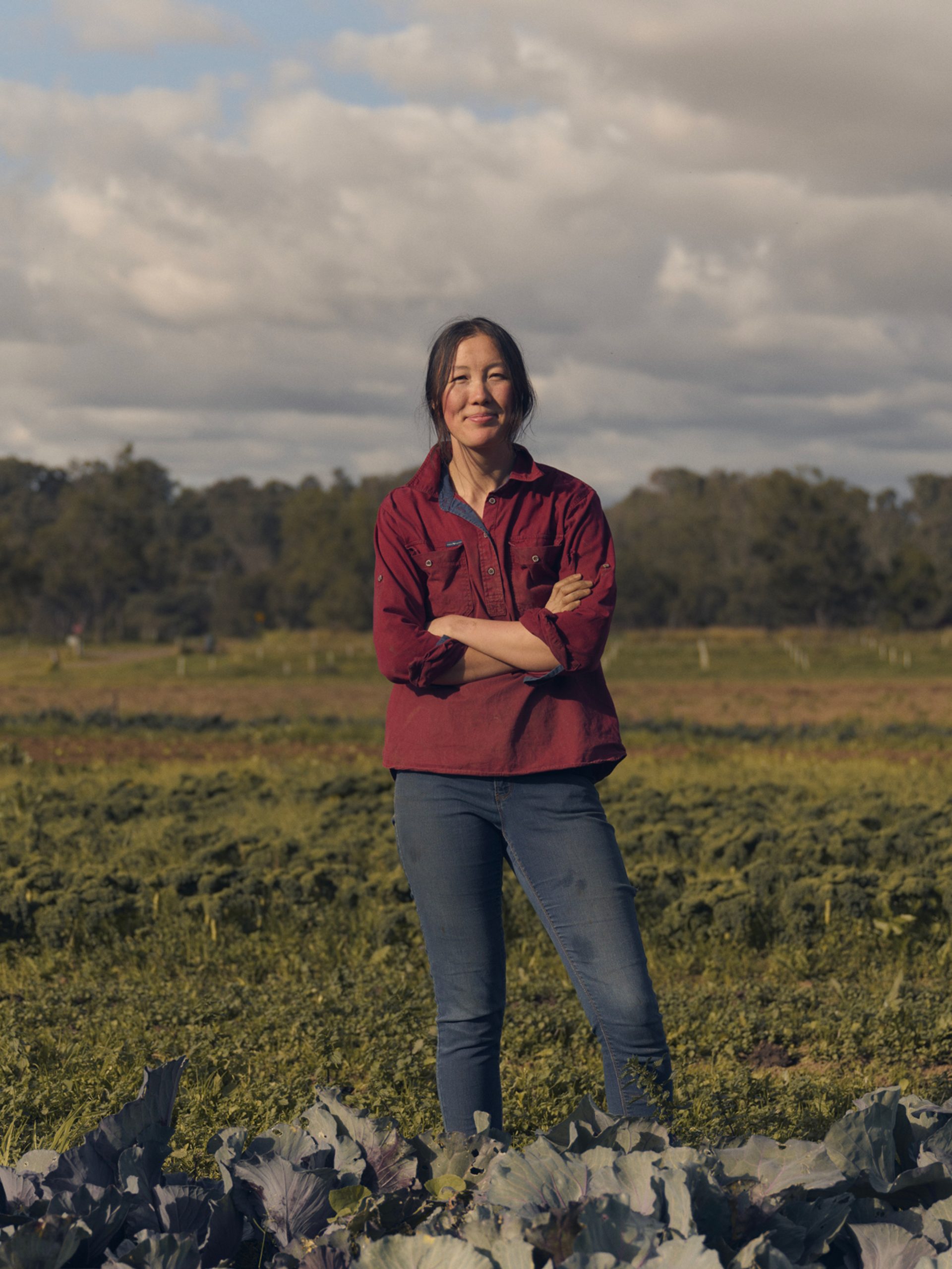 A woman stands confidently in a vibrant green field, surrounded by rows of leafy crops. She is wearing a red button-up shirt and jeans, with her arms crossed as she smiles at the camera. The background features expansive farmland under a partly cloudy sky, with a distant line of trees framing the horizon.