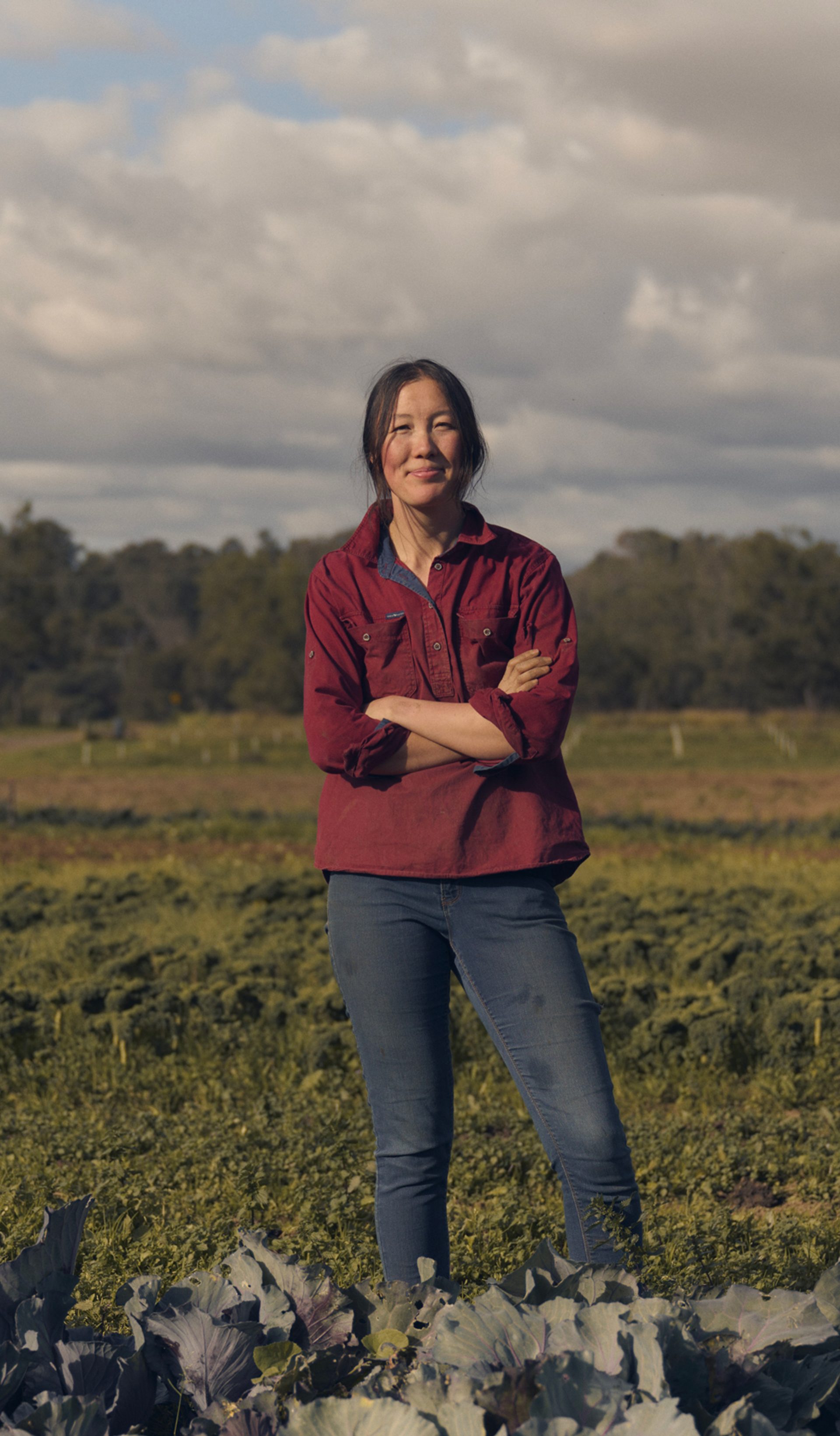 A woman stands confidently in a vibrant green field, surrounded by rows of leafy crops. She is wearing a red button-up shirt and jeans, with her arms crossed as she smiles at the camera. The background features expansive farmland under a partly cloudy sky, with a distant line of trees framing the horizon.