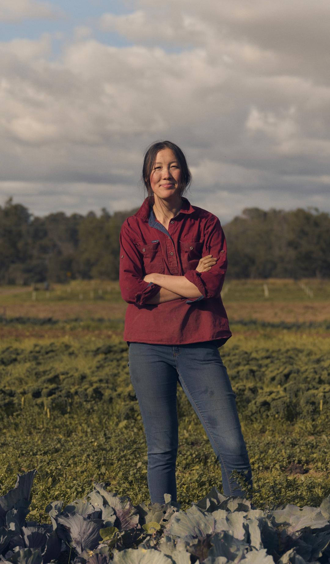 A woman stands confidently in a vibrant green field, surrounded by rows of leafy crops. She is wearing a red button-up shirt and jeans, with her arms crossed as she smiles at the camera. The background features expansive farmland under a partly cloudy sky, with a distant line of trees framing the horizon.