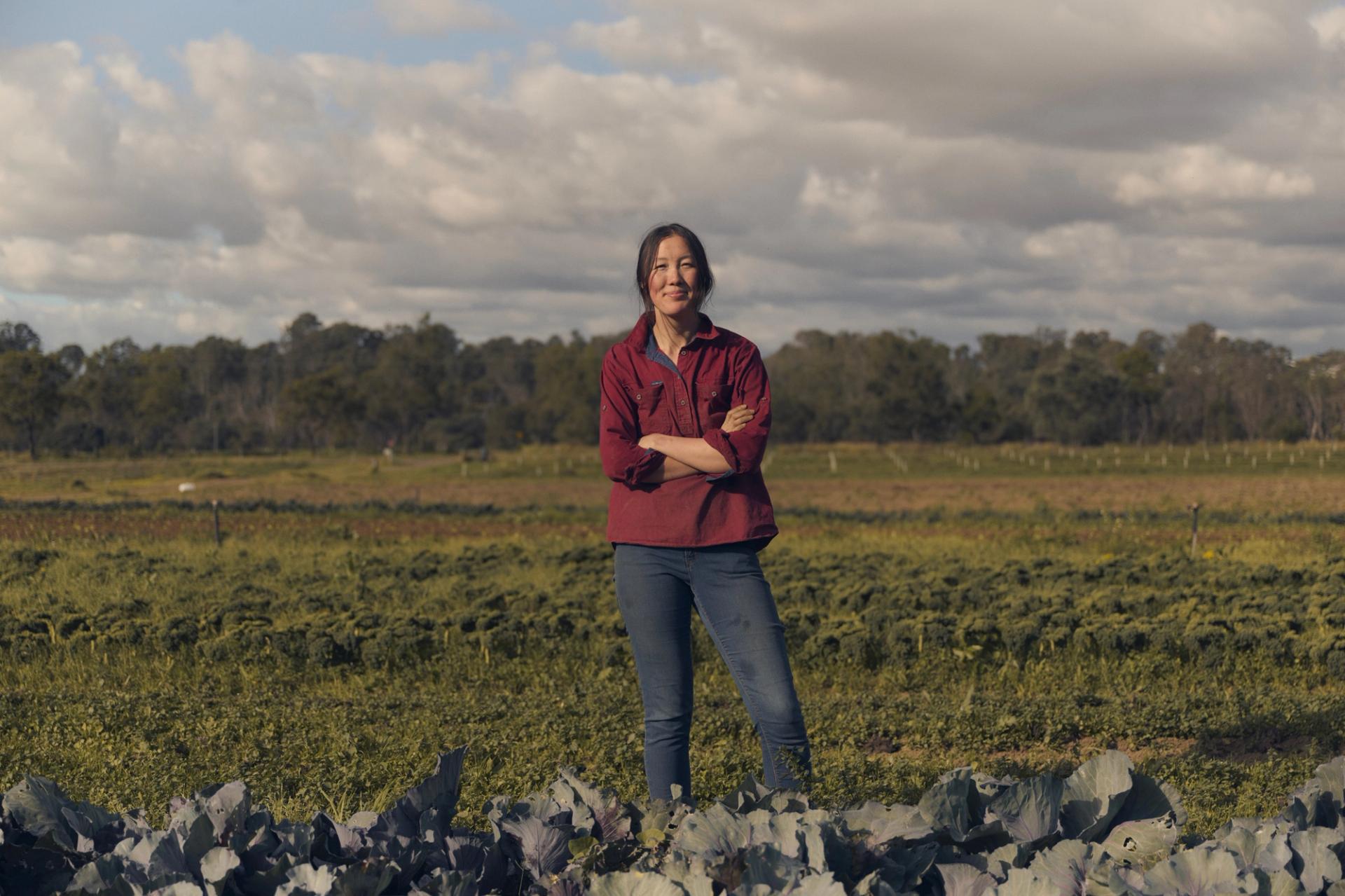 A woman stands confidently in a vibrant green field, surrounded by rows of leafy crops. She is wearing a red button-up shirt and jeans, with her arms crossed as she smiles at the camera. The background features expansive farmland under a partly cloudy sky, with a distant line of trees framing the horizon.