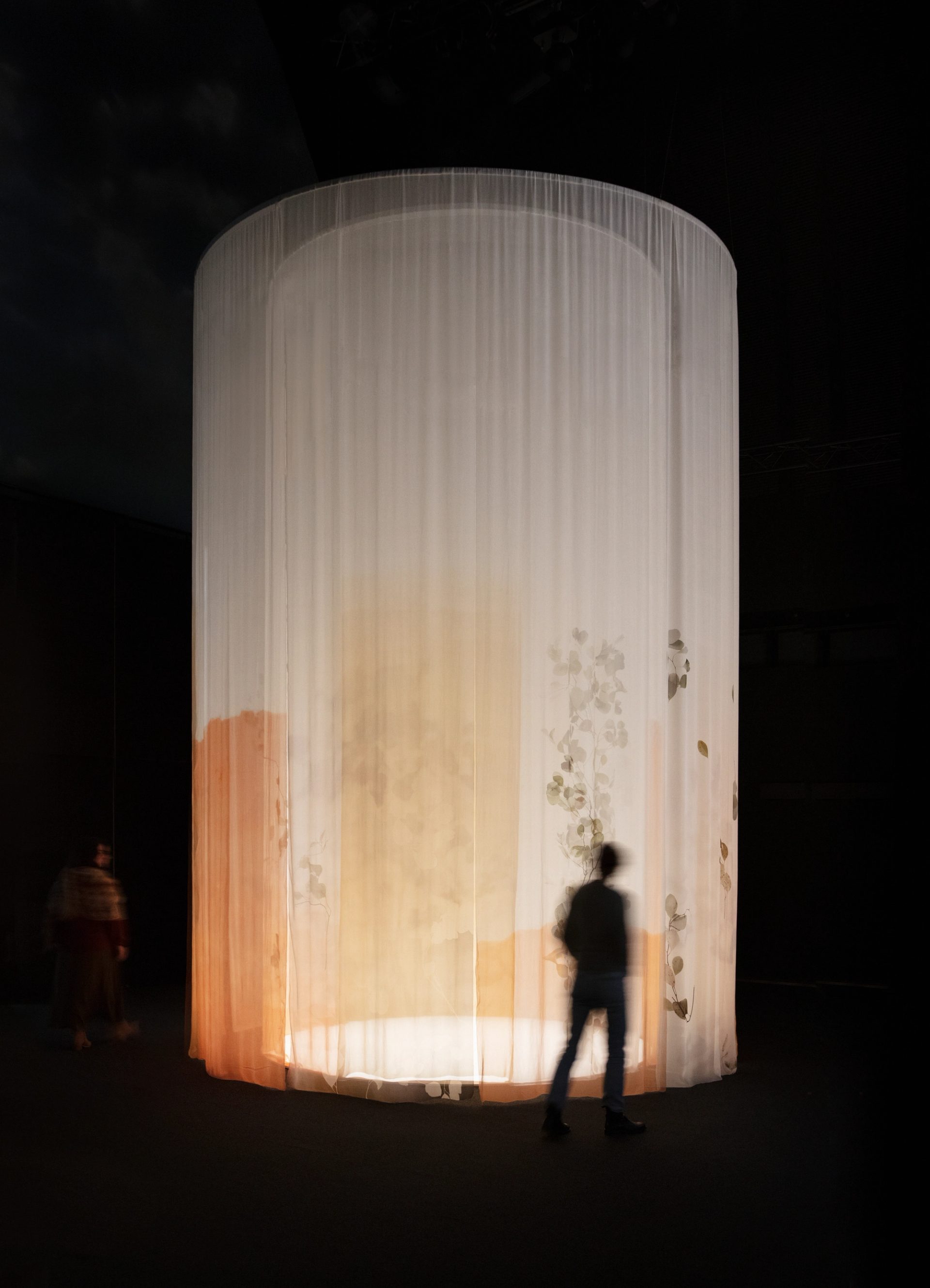 A tall white curtain hangs with patterns of foliage seen through the transparent fabric. A blurred silhouetted figures stands in front.